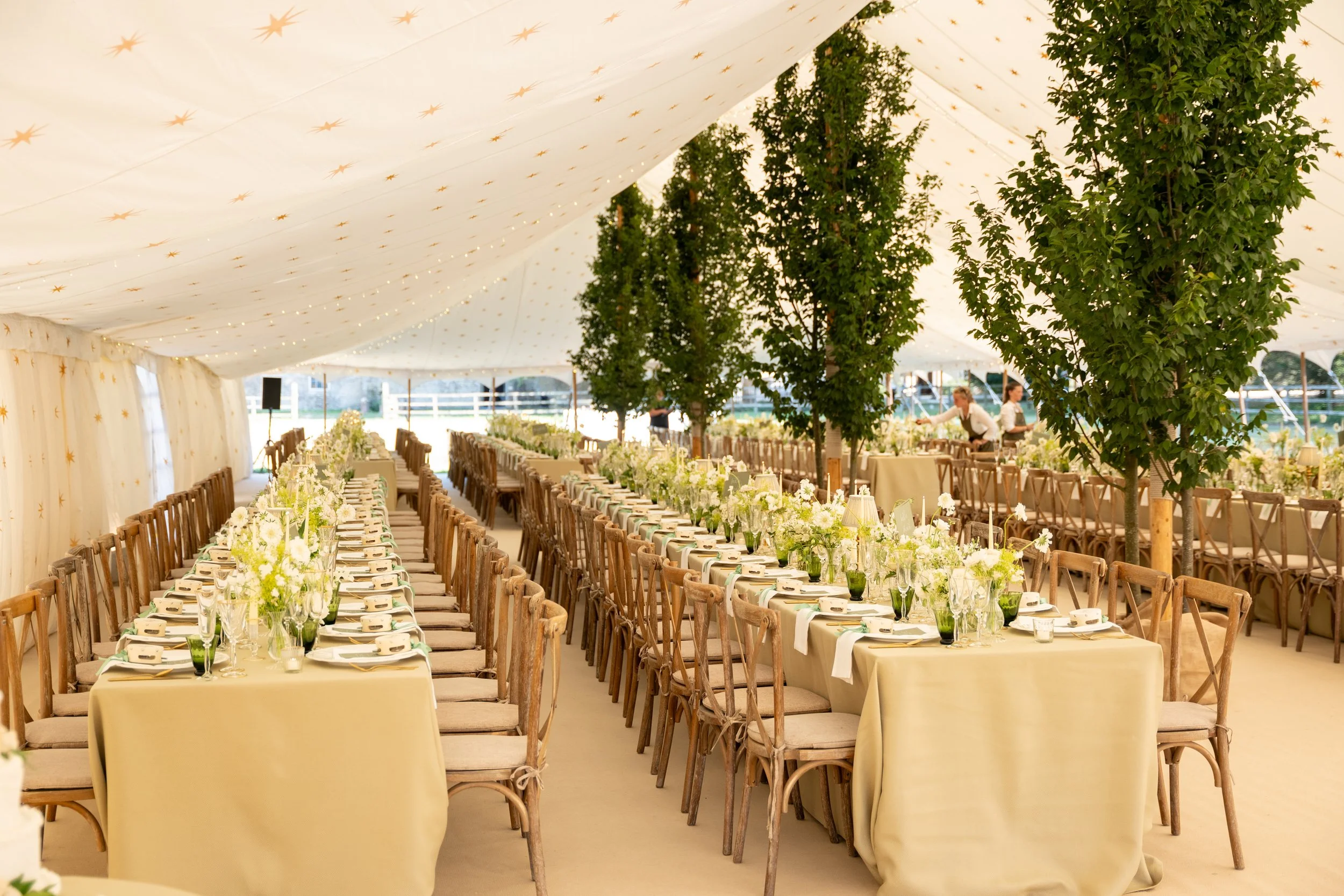 Long banquet tables decorated with white flowers and tableware set up inside a white tent with star patterns and green trees.