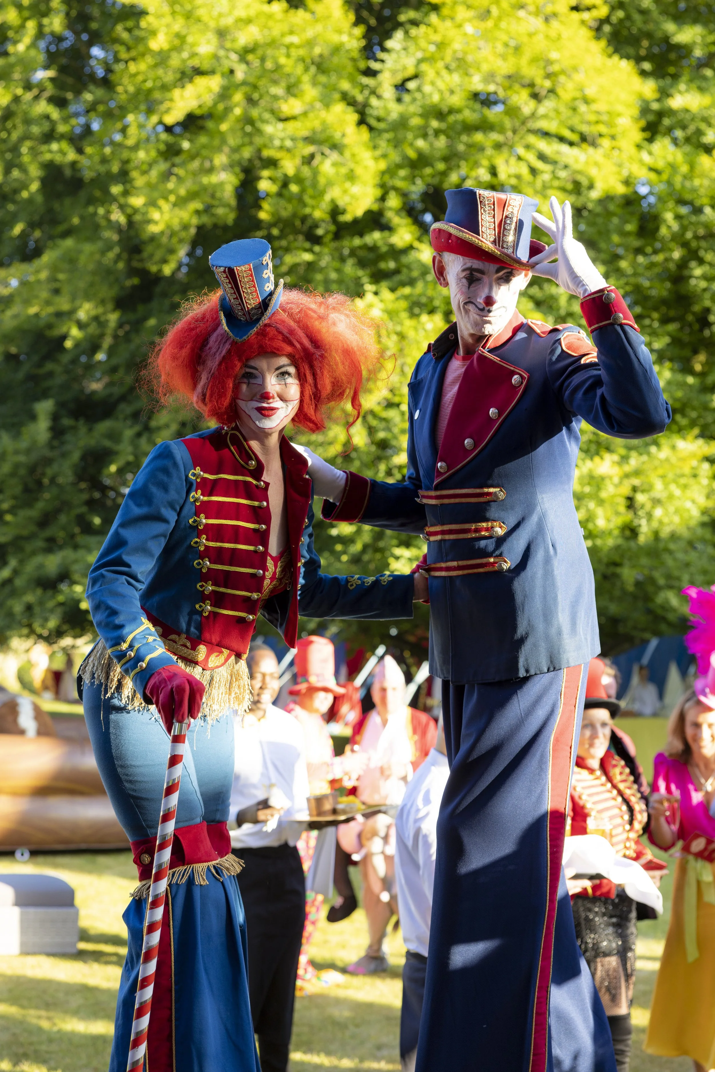 Two circus performers dressed as clowns in colorful costumes, standing outdoors with a green leafy background and other people in the distance.