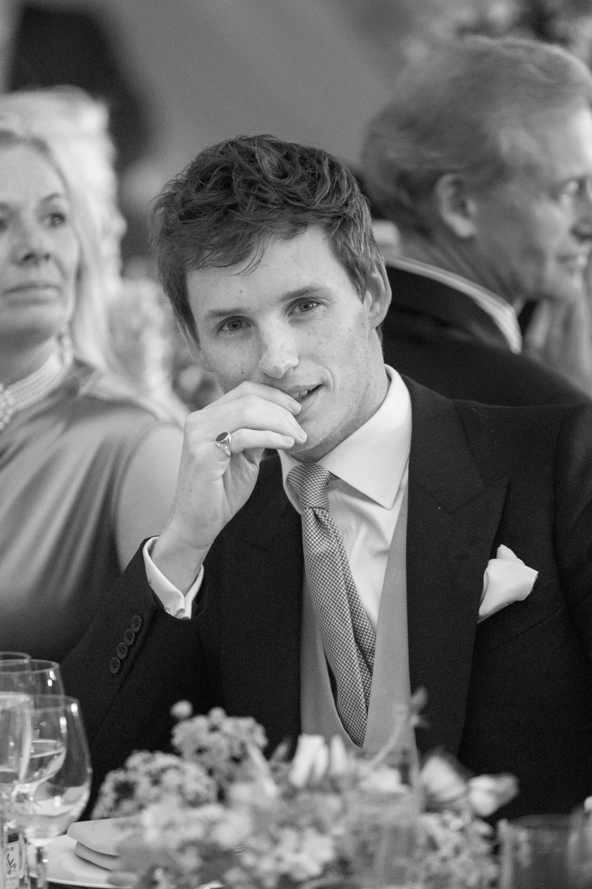 A young man in a suit sitting at a formal event, looking at the camera with his hand near his mouth, surrounded by older adults and table settings.
