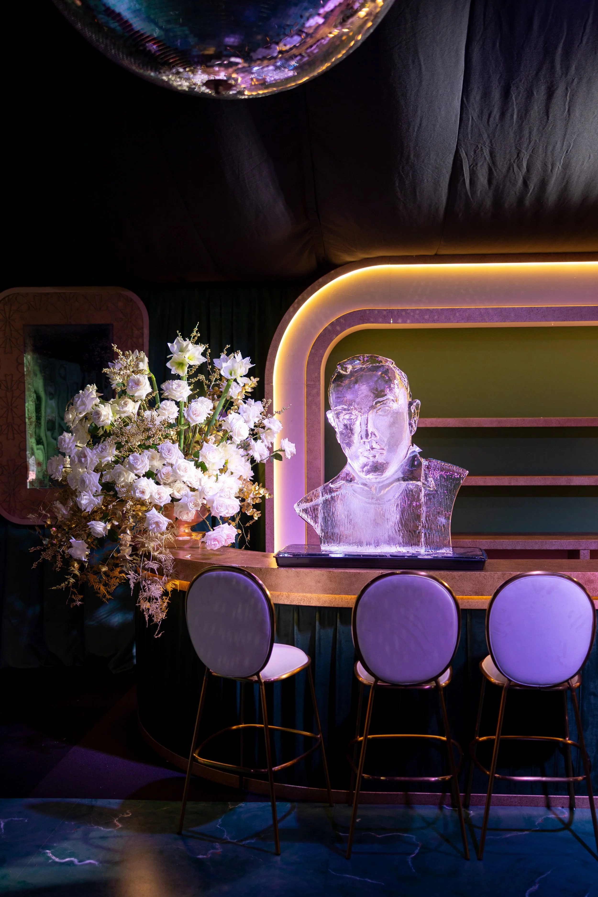 An ice sculpture of a person's bust on a bar counter, with a large bouquet of white flowers beside it, and a neon-lit decorative background.