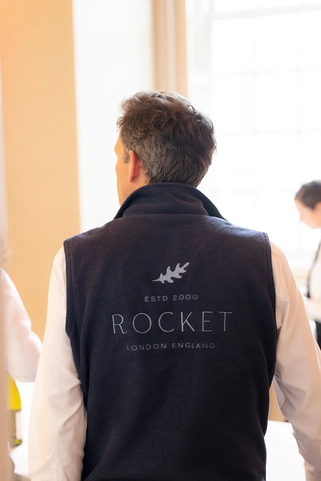 Back view of a man wearing a black vest with the Rocket logo and text, standing indoors near a window.