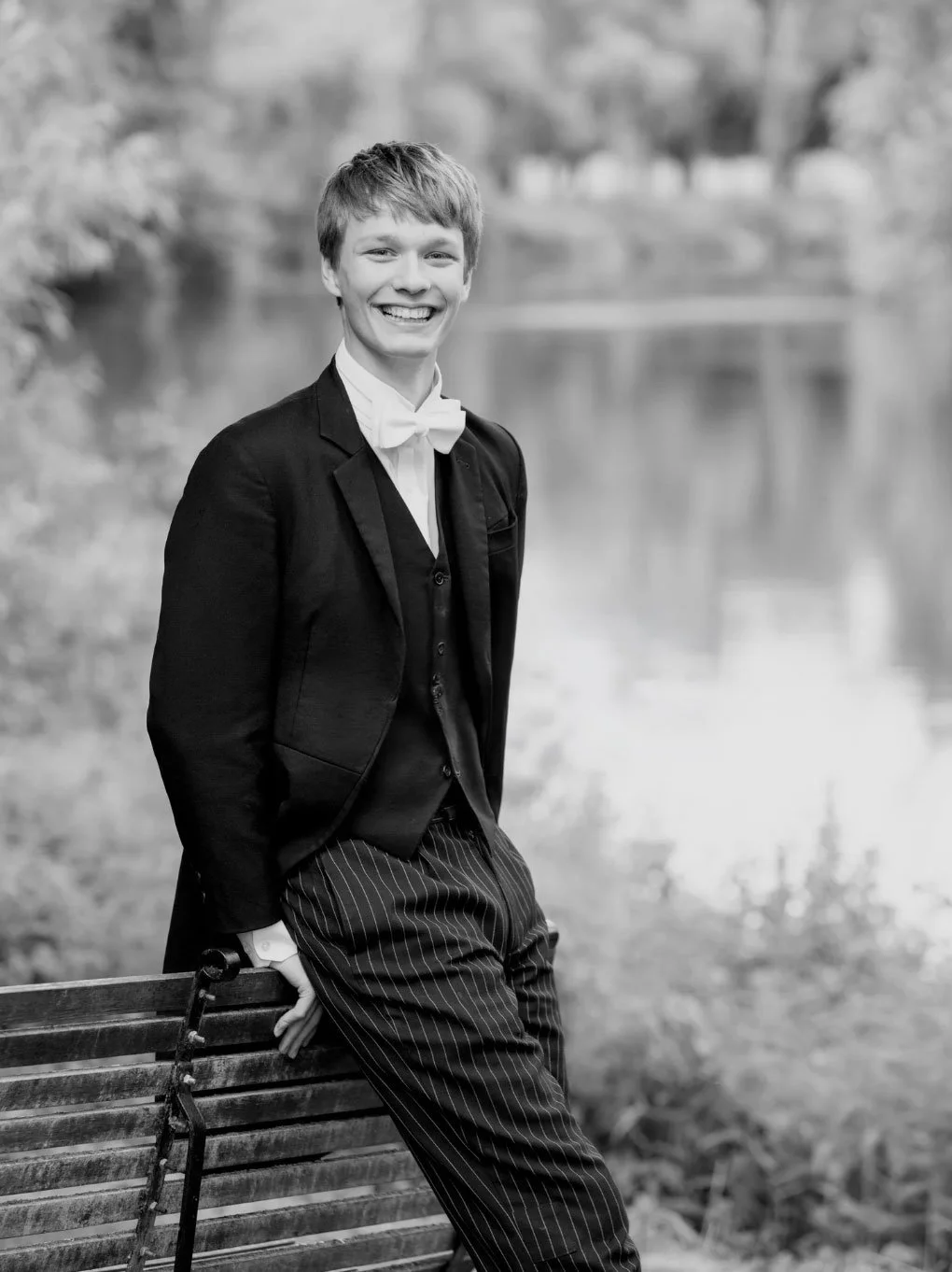 A young man dressed in formal attire, including a tuxedo with a bow tie, standing outdoors next to a bench near a river, smiling at the camera.