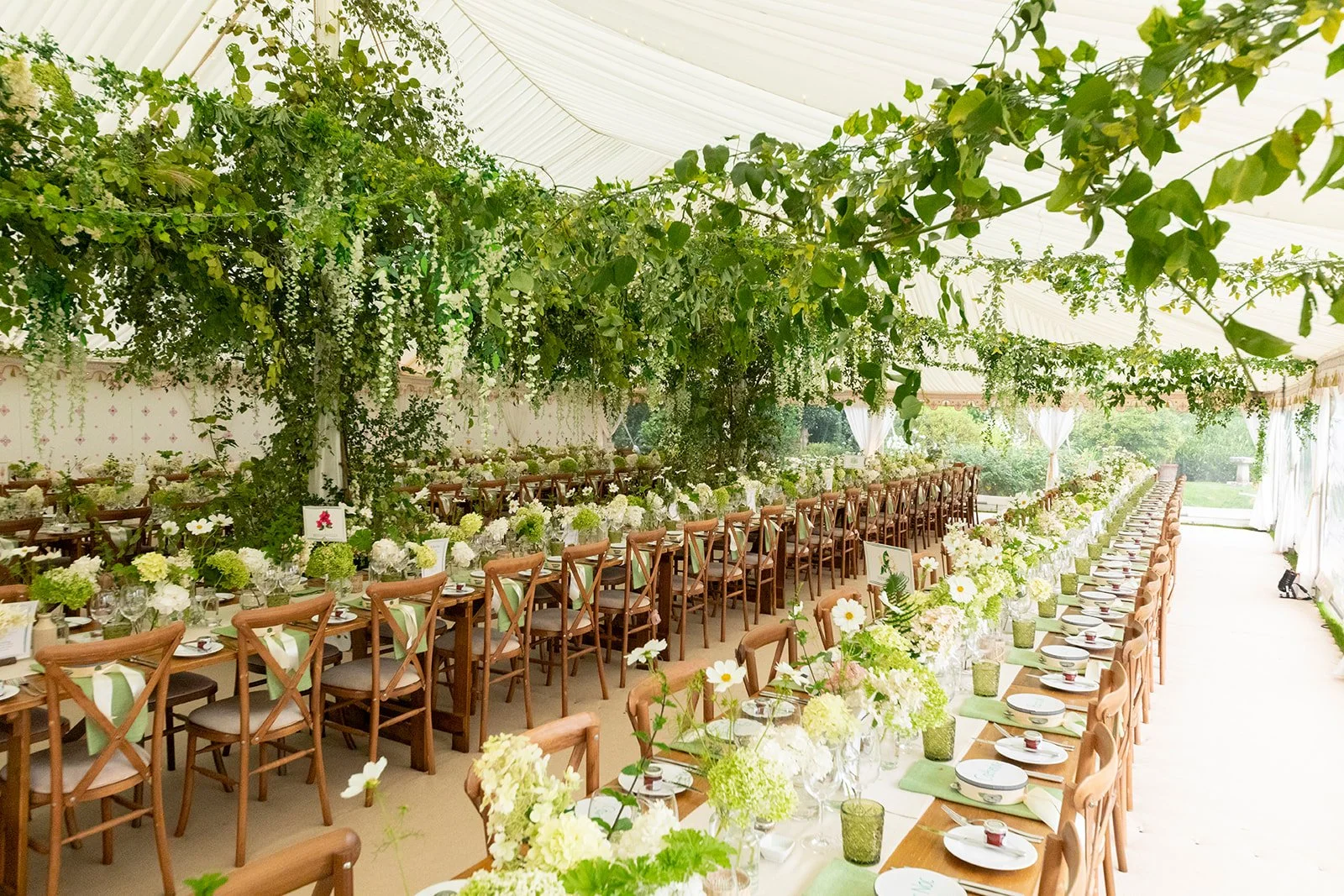 Long banquet tables decorated with white and green floral arrangements, set with plates, glasses, and cutlery inside a white tent with hanging greenery and trees outside.