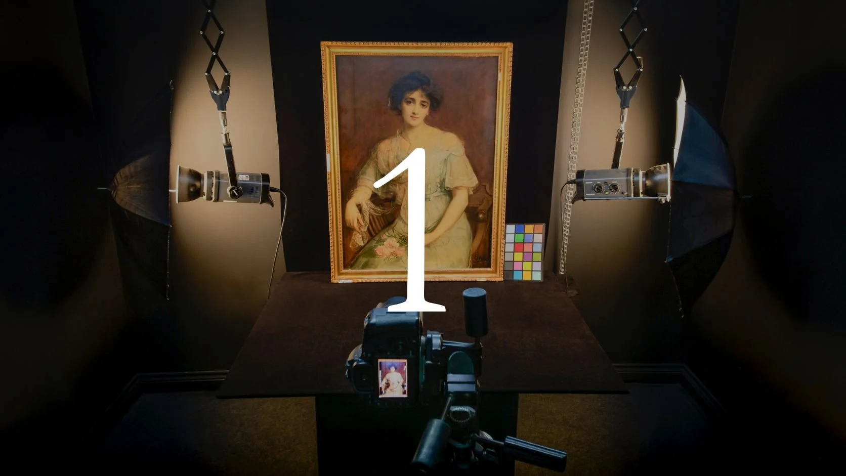 Photography setup with two studio lights illuminating a framed portrait of a woman, a color calibration chart, a camera on a tripod taking a picture of the portrait, and a color reference card, inside a dark room.