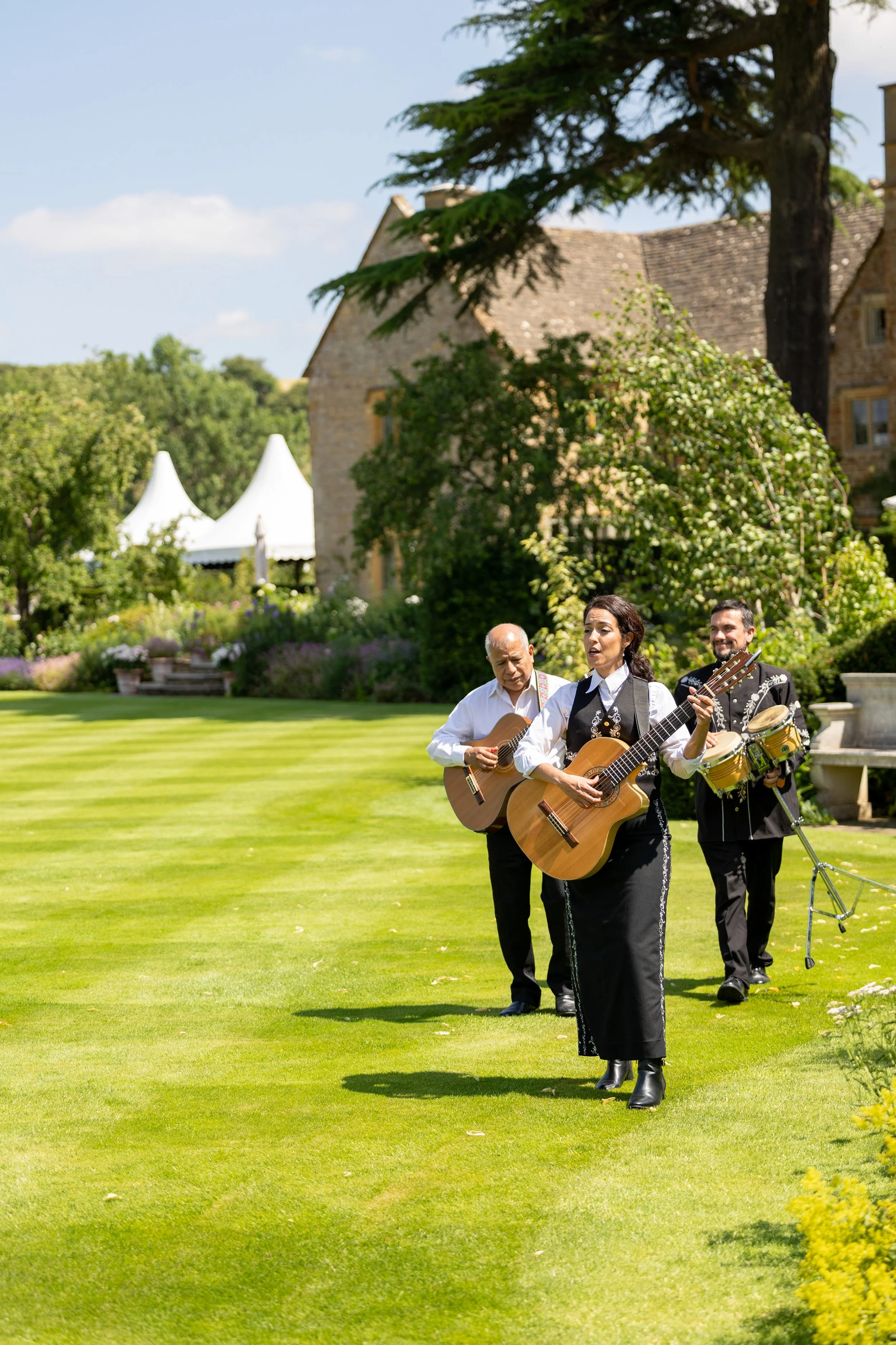 A group of three musicians playing guitars and percussion outdoors on a sunny day, with a large tree, a stone building, and a garden in the background.