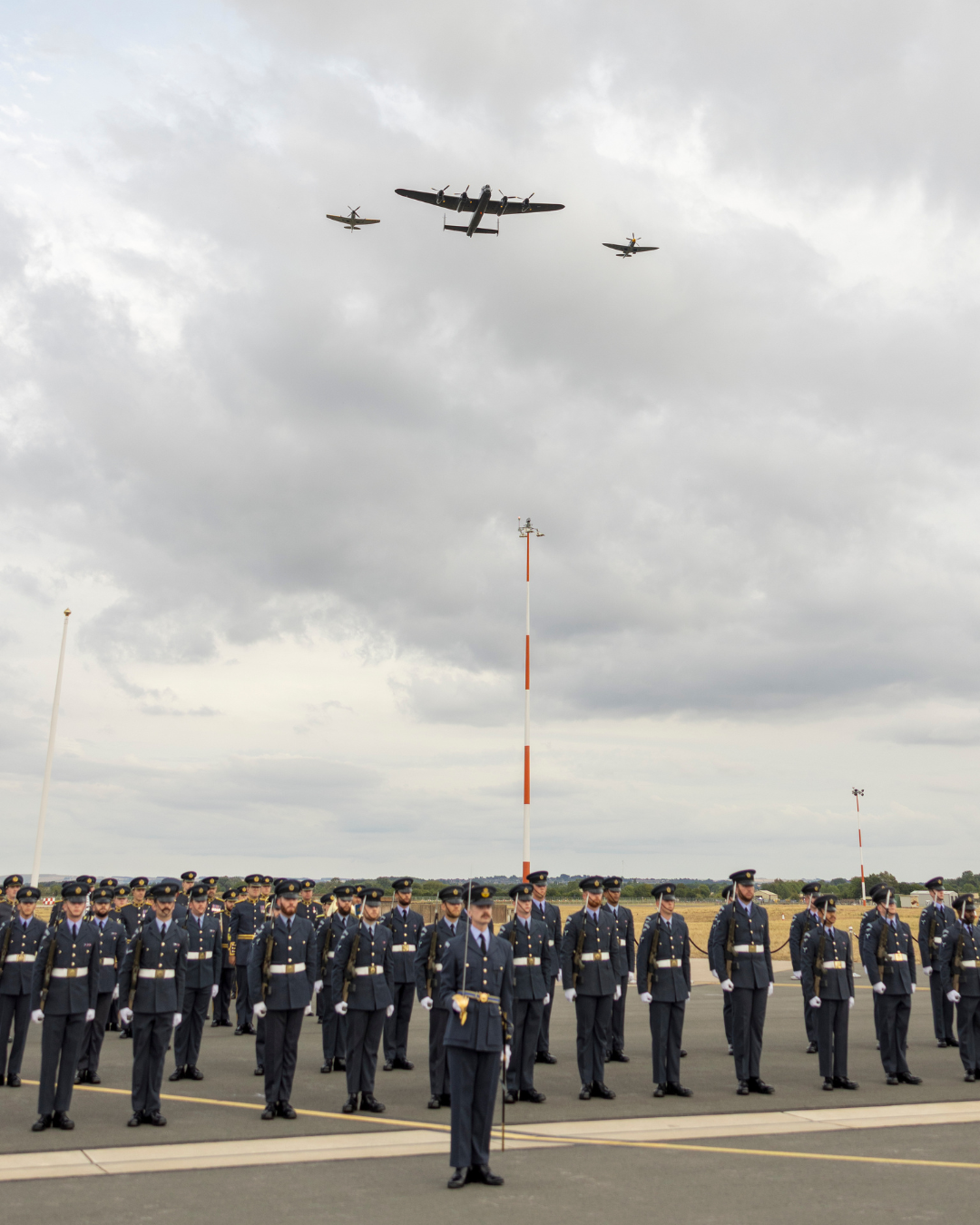 Military personnel standing in formation at an airfield with planes flying overhead.
