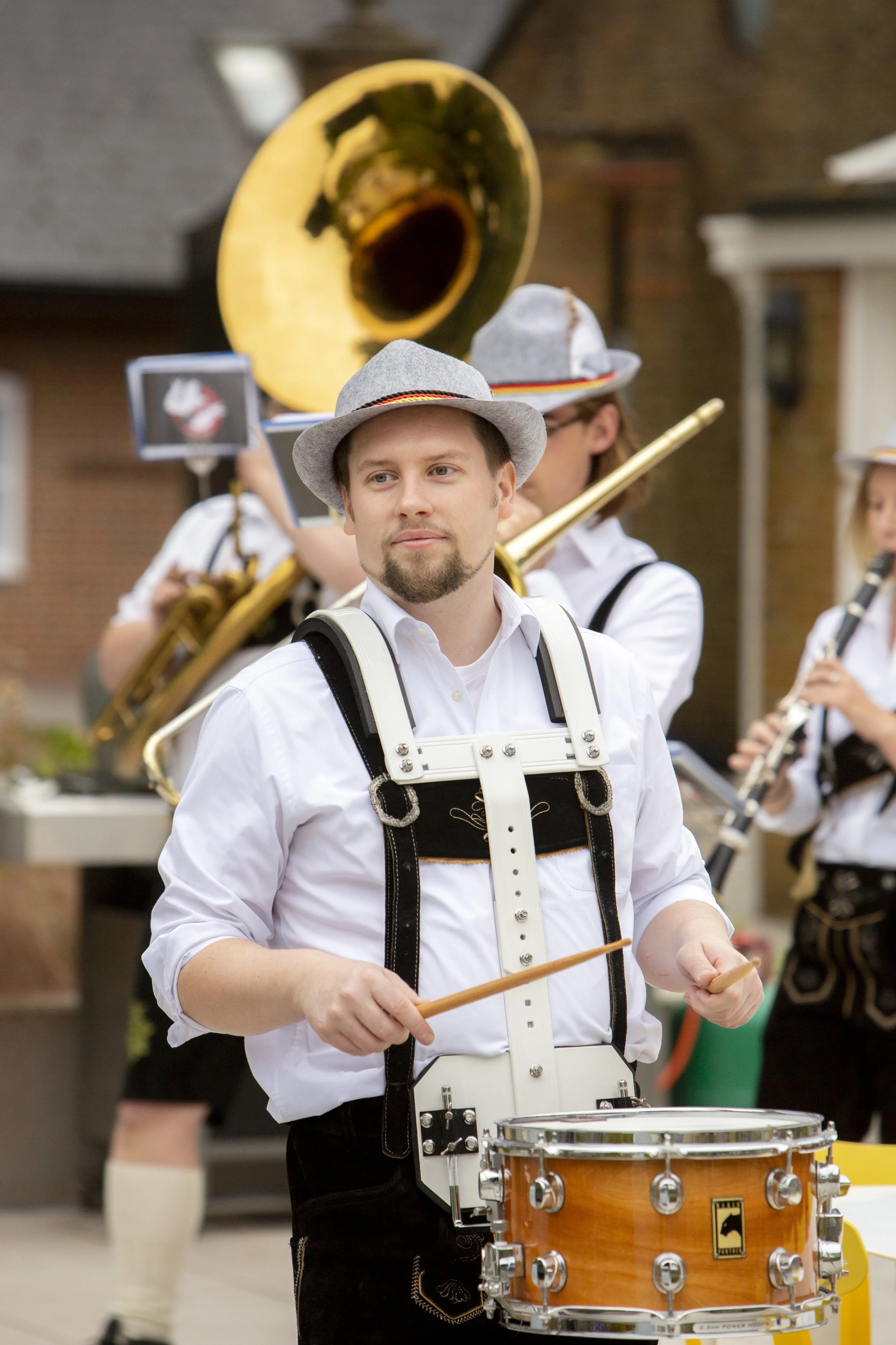 A man dressed in traditional Alpine attire playing a drum at an outdoor event with a band in the background, including a sousaphone and a clarinet, wearing hats and white shirts.