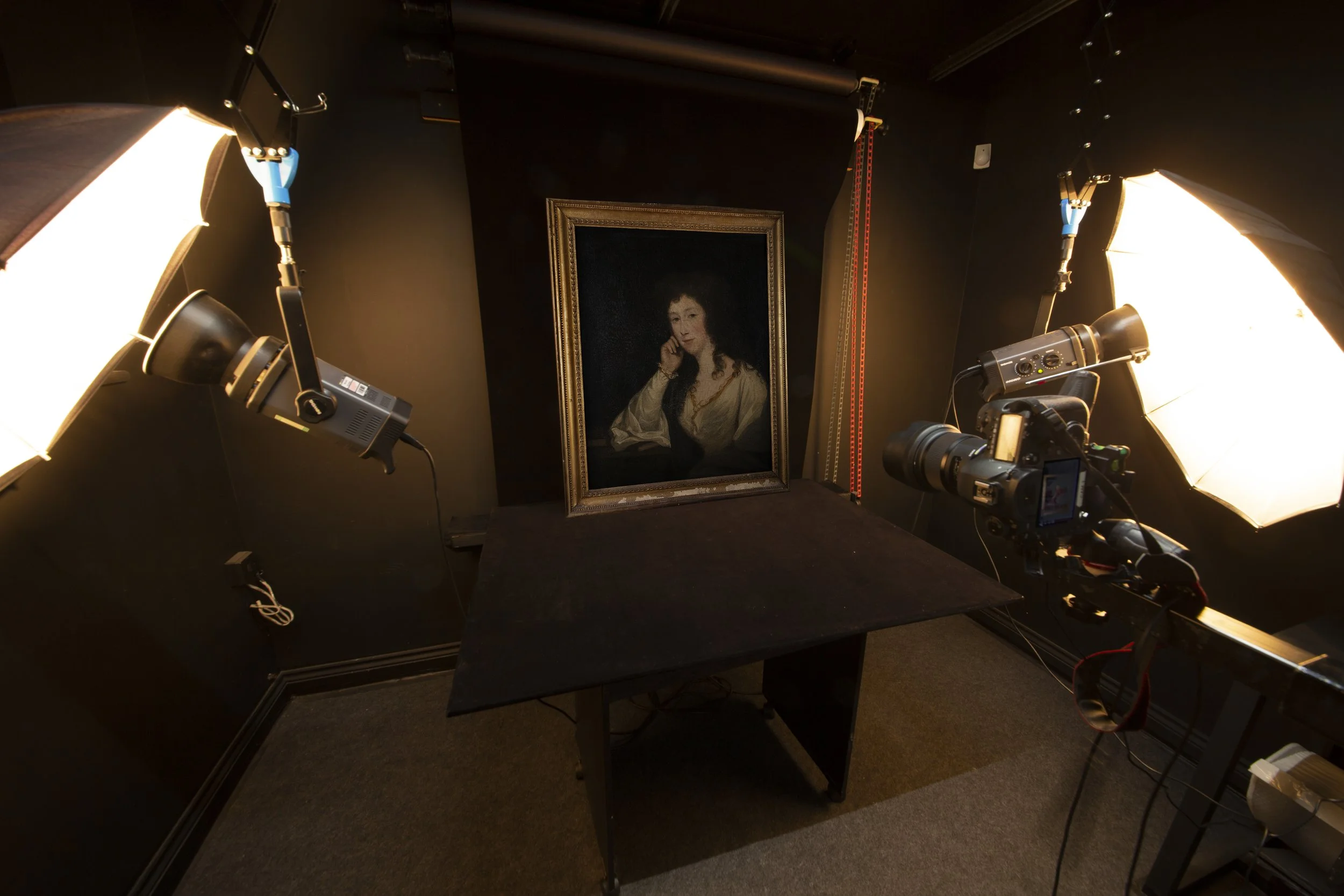 Photography studio setup with professional lighting equipment focused on a framed portrait of a woman with dark, curly hair, wearing a cream-colored blouse. Camera on tripod in the foreground, dark walls, and black table surface.