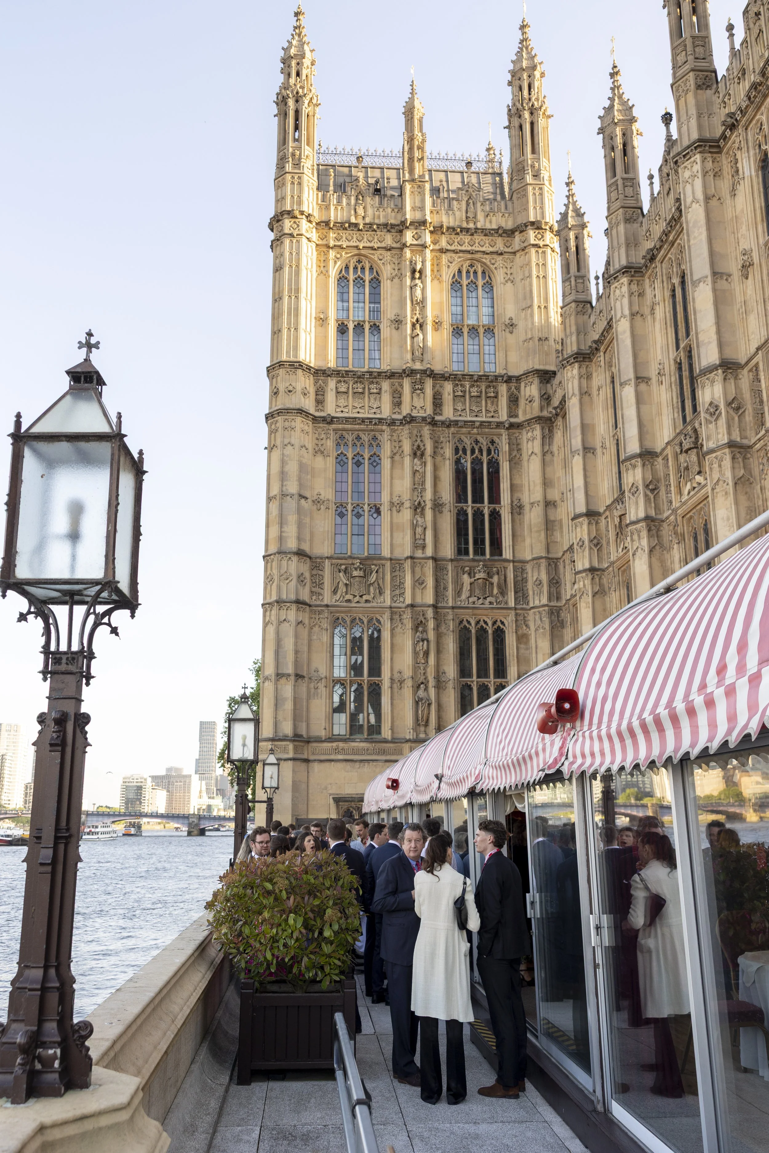 People in formal attire gathered outside a building with Gothic architectural features, near a river lined with urban buildings, in London.