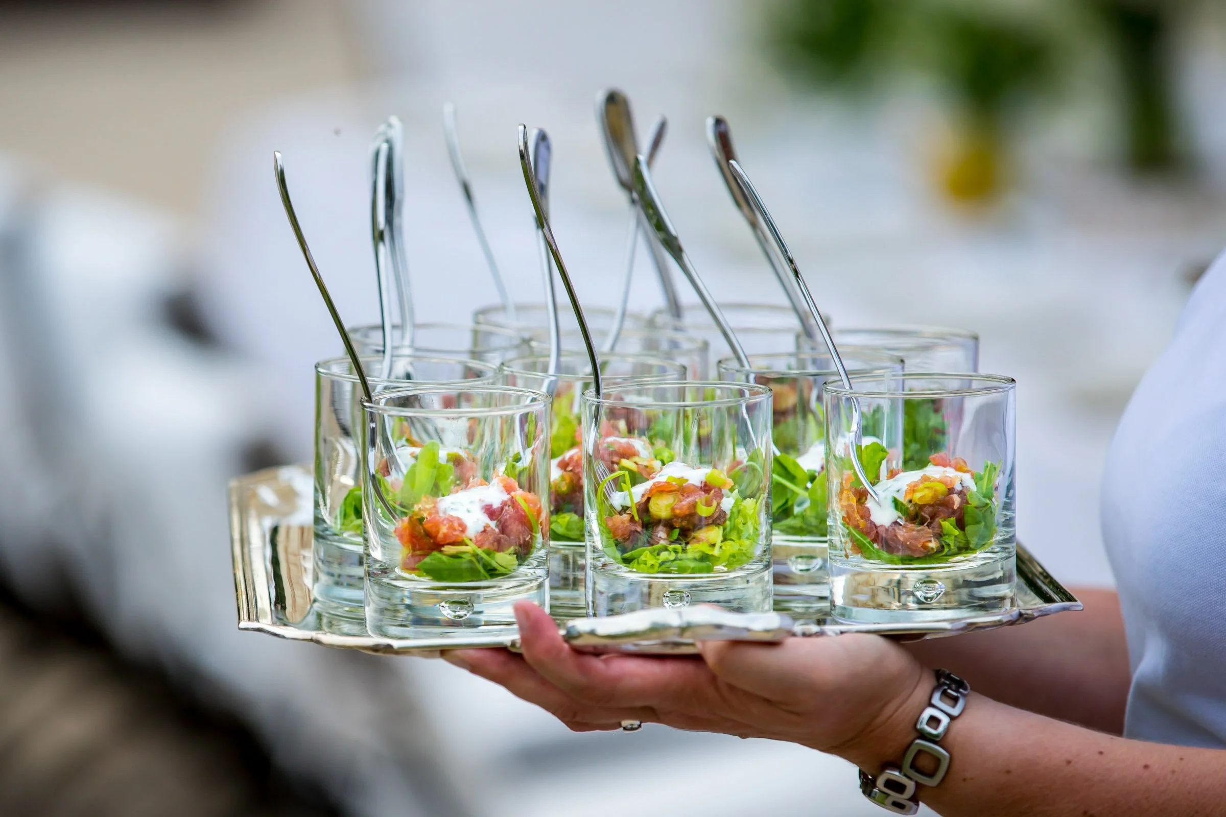 A person holding a silver tray with small glass cups filled with salad, each with a spoon.