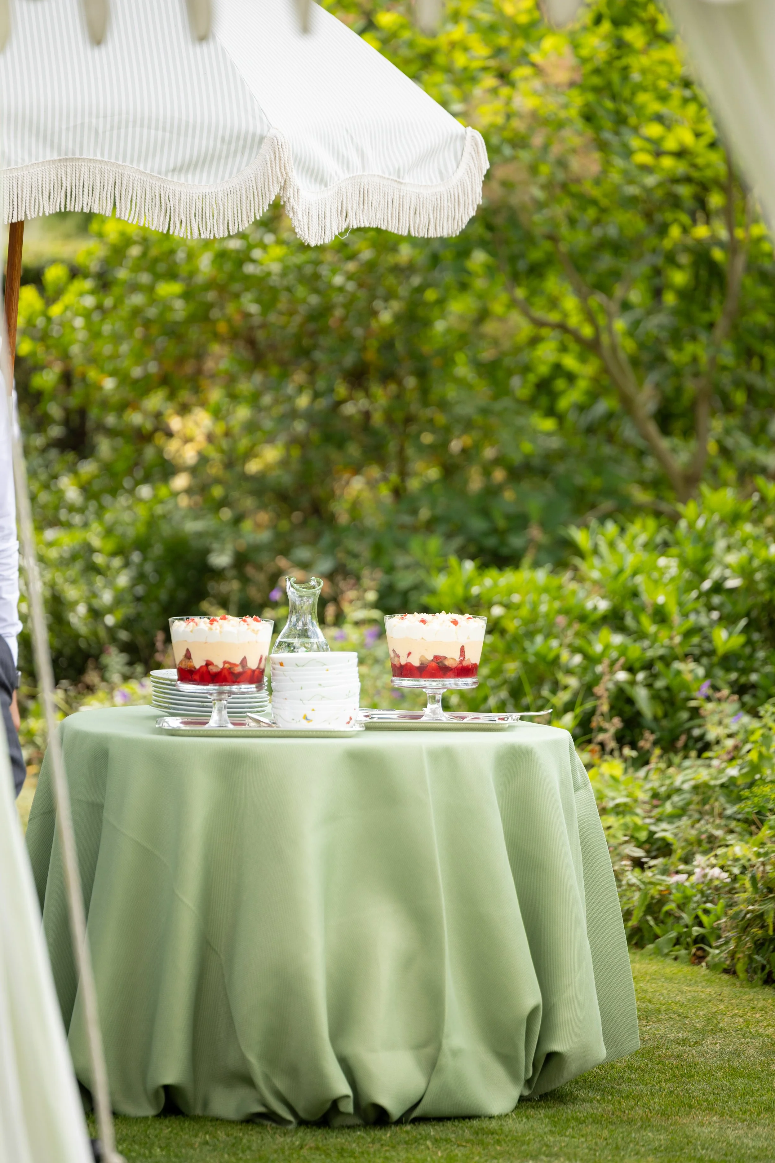 A table with a light green tablecloth set outdoors under a white umbrella, with two strawberry and cream desserts and a glass pitcher in the center, surrounded by greenery.