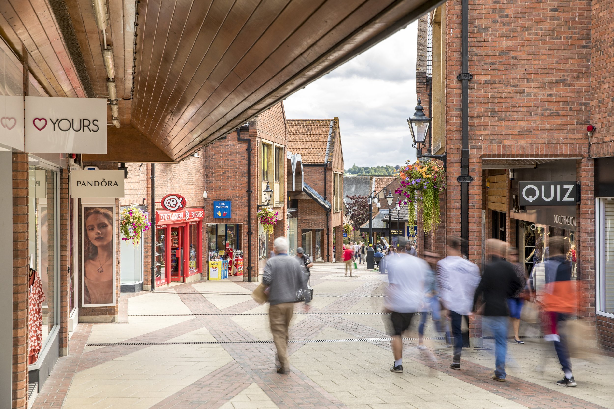 A busy outdoor shopping street with shops on both sides, hanging flower baskets, and people walking, some appearing blurred due to motion.