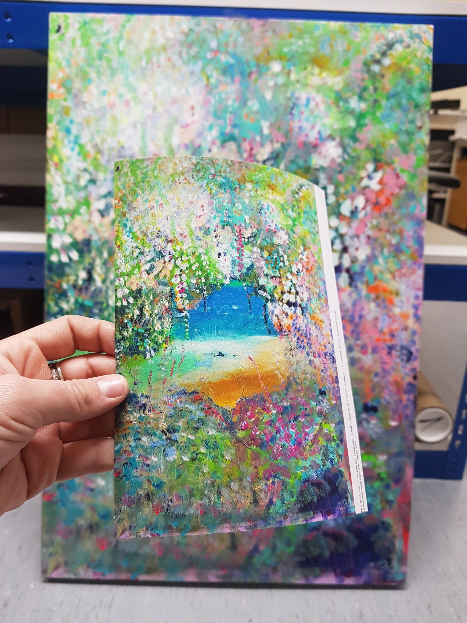 Hand holding a colorful painting of a garden with a waterfall in the background, viewed in front of a larger, similarly colorful artwork.