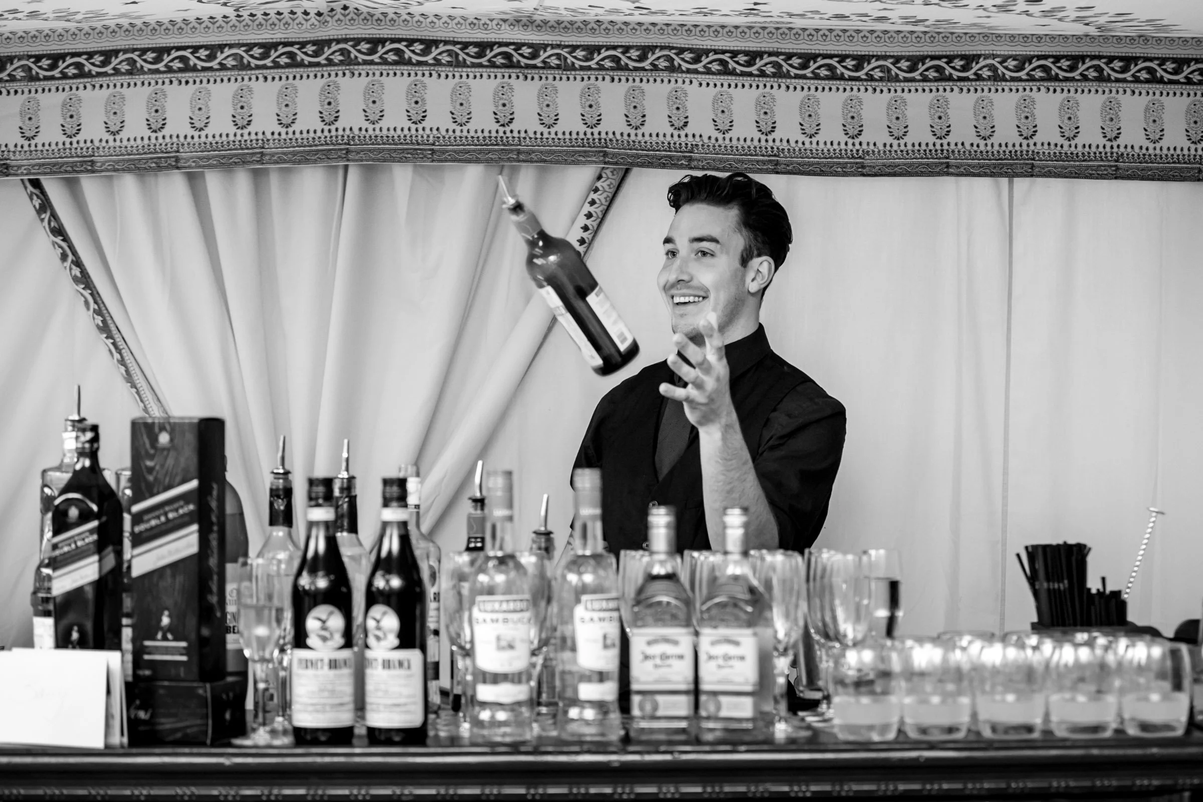 A smiling bartender pours a drink at a bar with various bottles and glasses in front of a curtain.