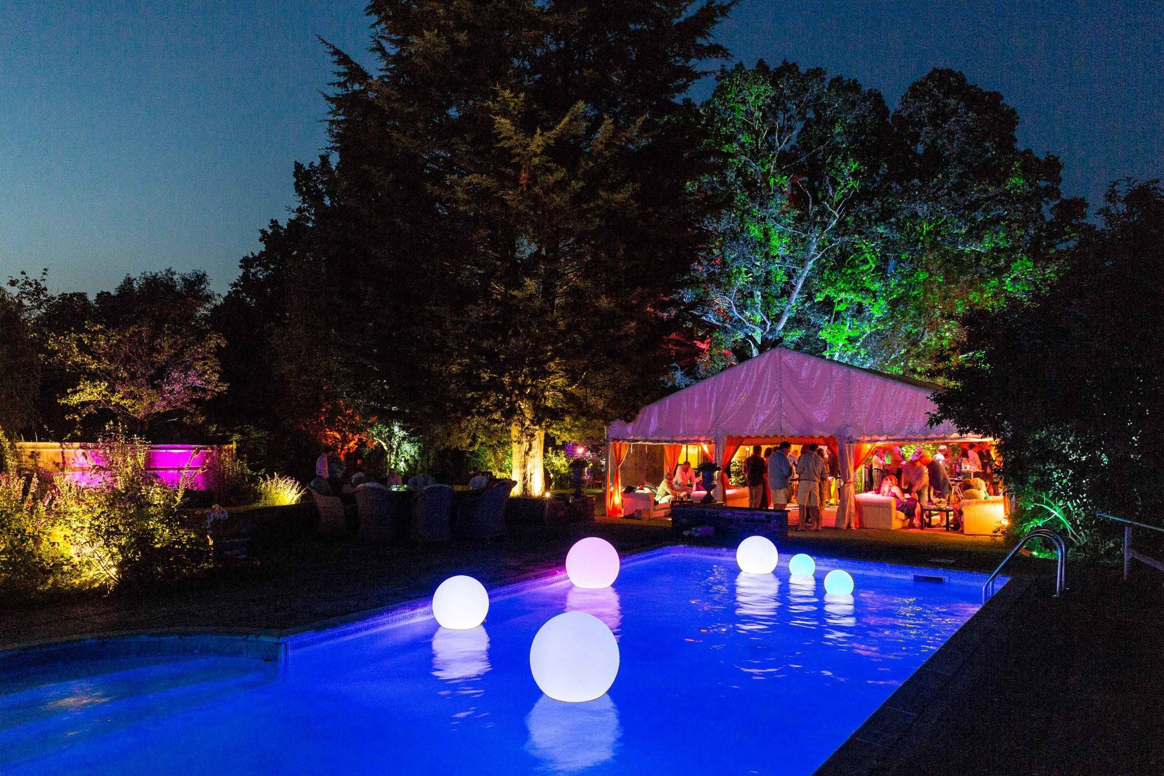 Nighttime outdoor party with illuminated pool, colorful trees, and a lit tent with guests socializing and relaxing.