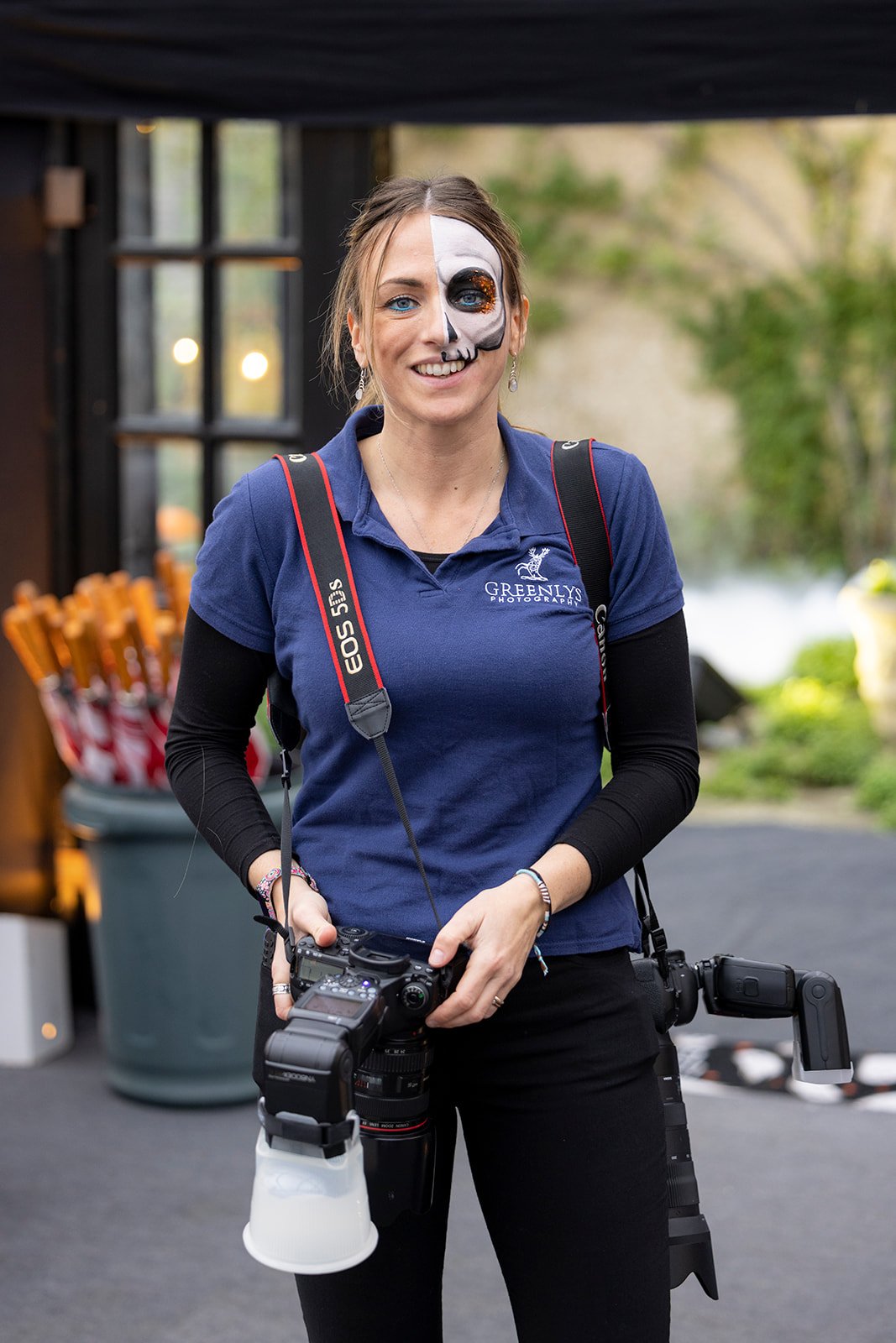 A woman with half of her face painted as a skeleton smiling outdoors, holding a professional camera, wearing a blue polo shirt with a camera company logo, and a black long sleeve shirt underneath.