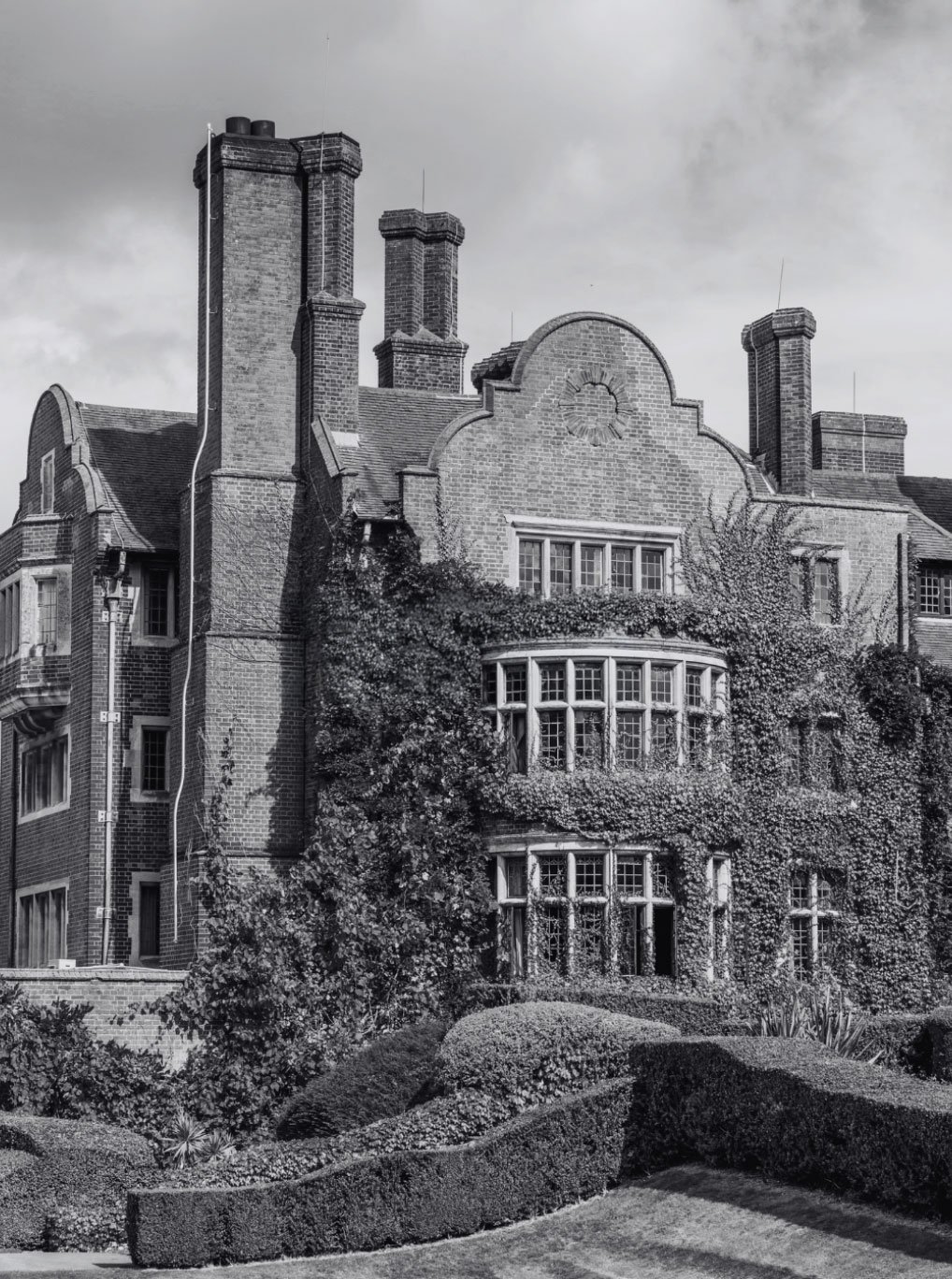 A large, historic brick house covered in climbing ivy, with multiple chimneys and bay windows, surrounded by well-manicured bushes and landscaping.