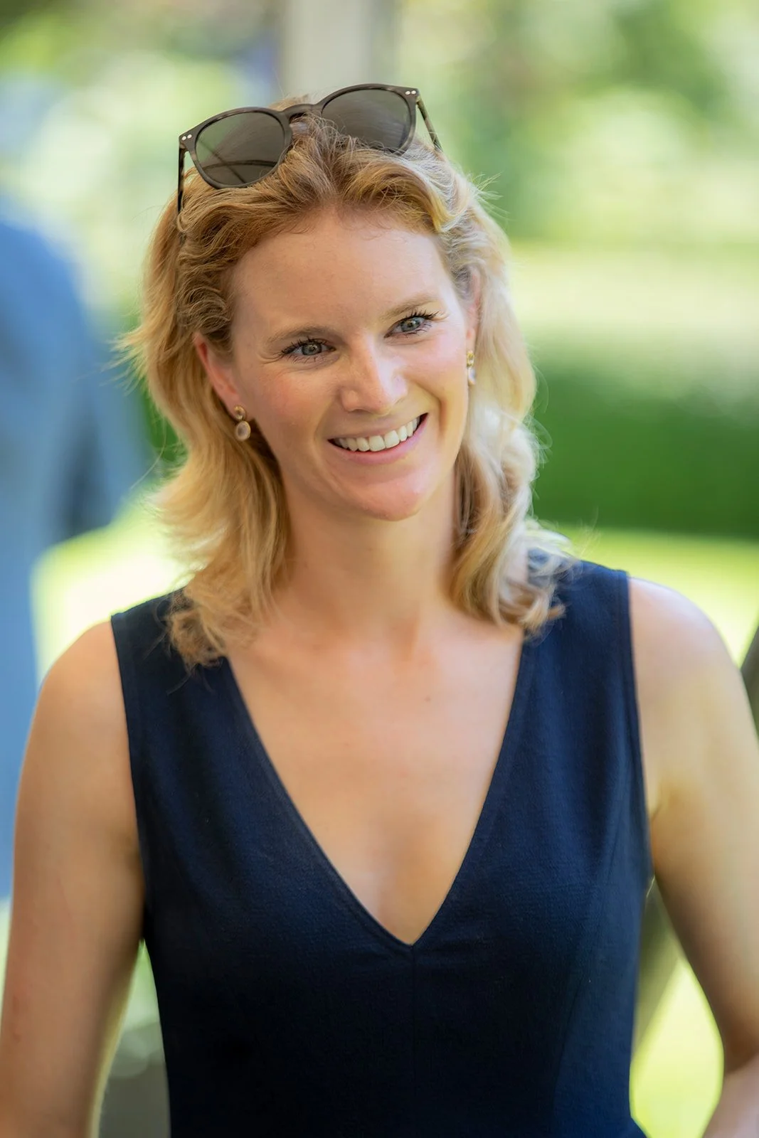 A smiling woman with blonde hair, wearing a black sleeveless dress, earrings, and sunglasses on her head, standing outdoors with trees and greenery in the background.