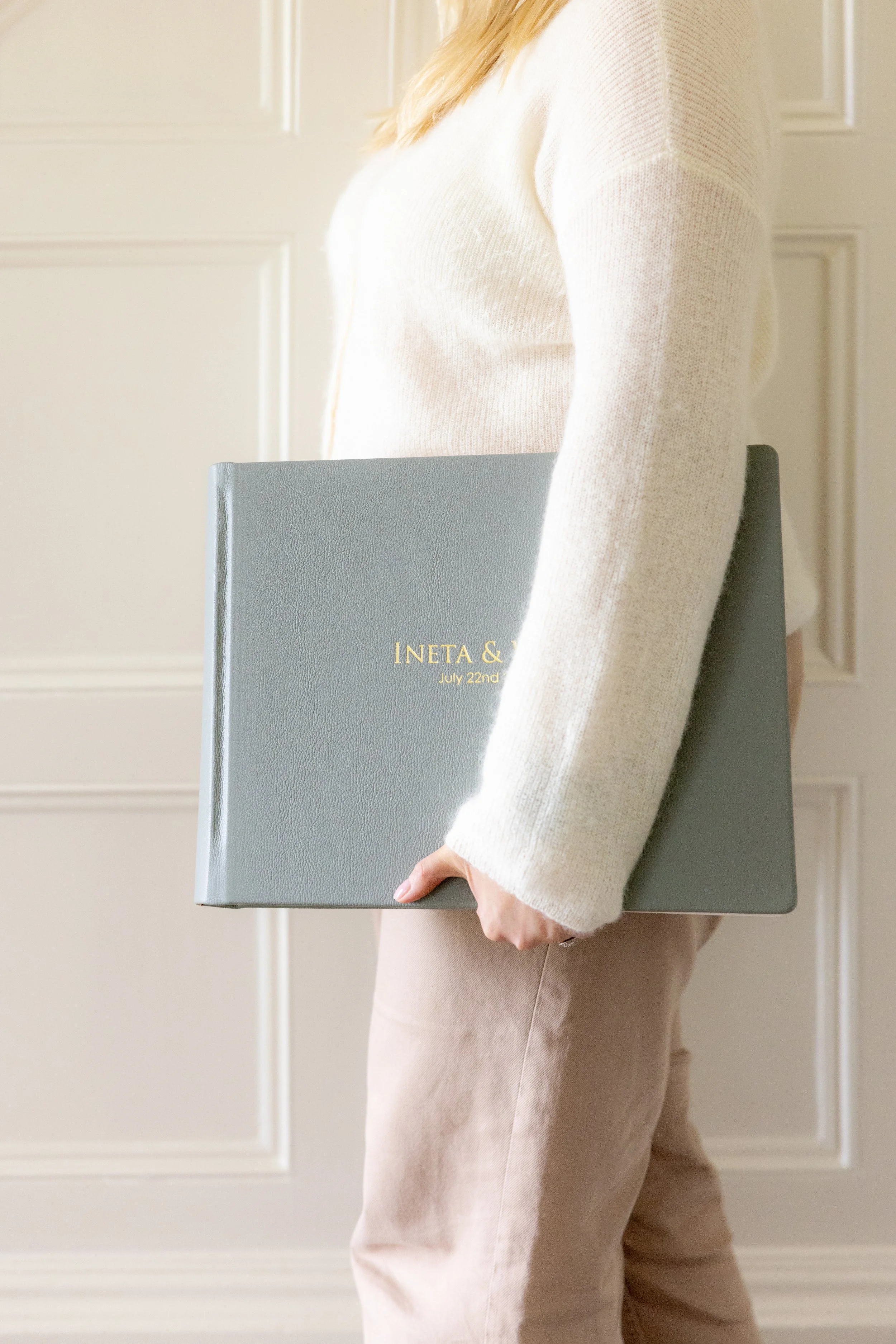 Person holding a gray portfolio with gold text that reads 'INETA &' and a date 'July 22nd'. The person is wearing a cream sweater and beige pants. The background consists of white paneled walls.