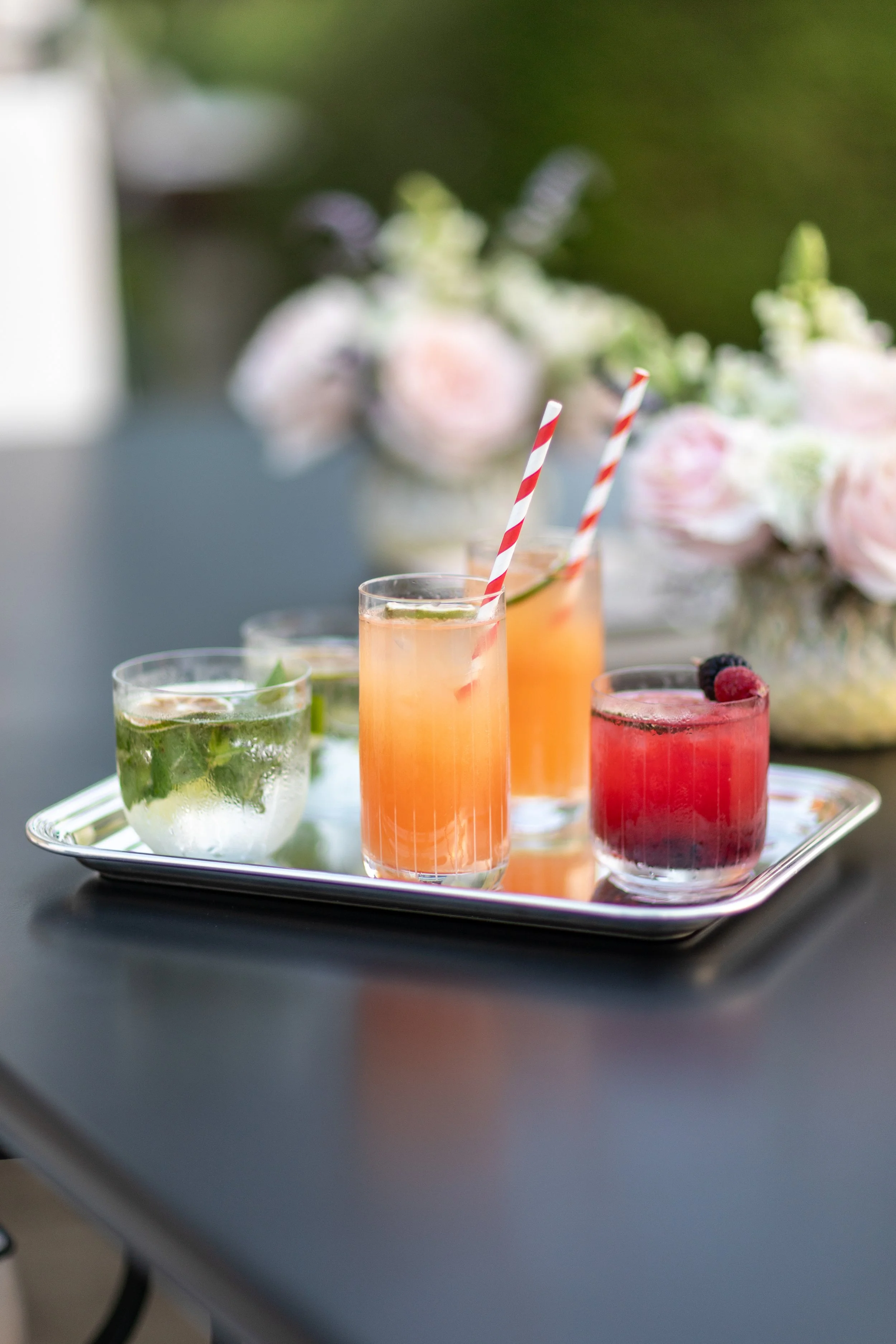 Four colorful cocktails on a silver tray with flowers in the background.