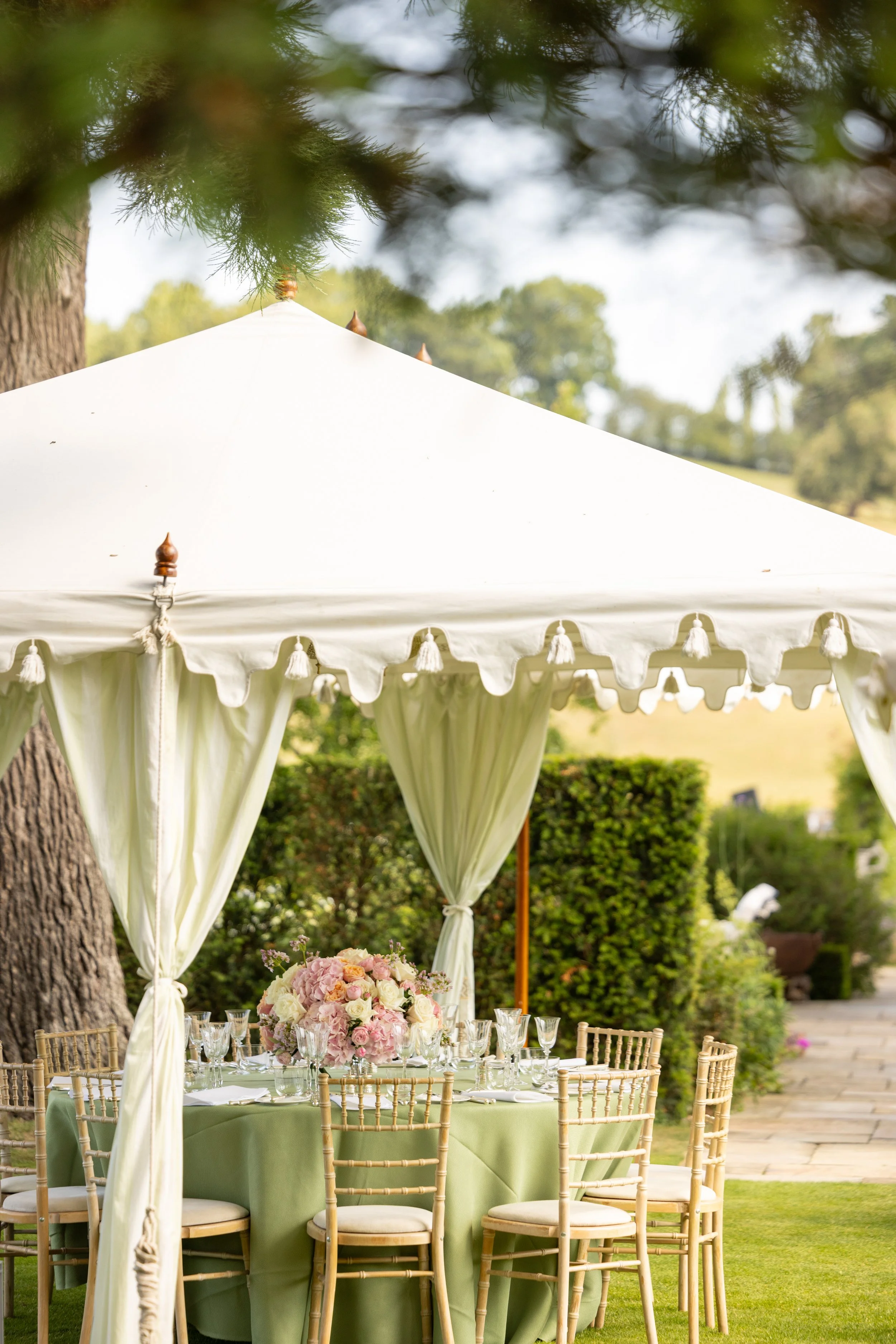 A round outdoor table set for a celebration under a white canopy tent with green tablecloths, floral centerpieces with pink and white flowers, surrounded by gold chairs, and lush green trees in the background.