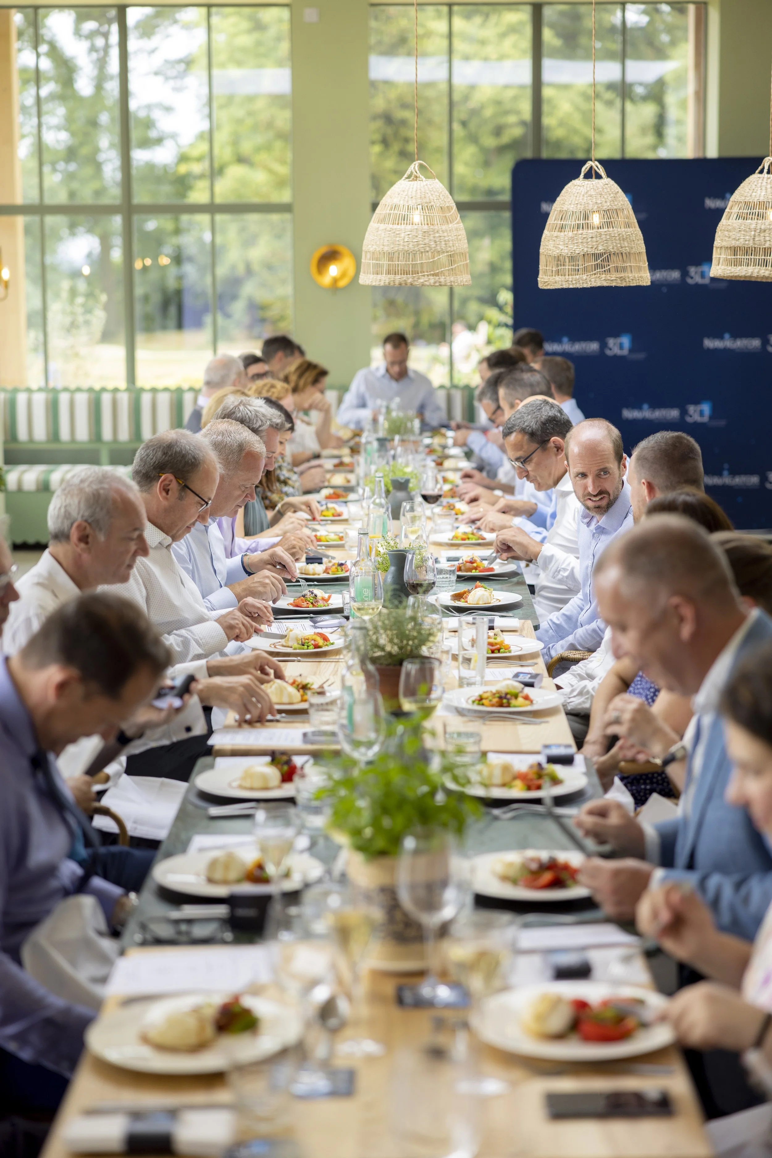 A large group of people are sitting at a long dining table in a well-lit room with large windows. They are having a meal, and there's greenery visible outside the windows. The table is set with plates of food, glasses, and small potted plants.