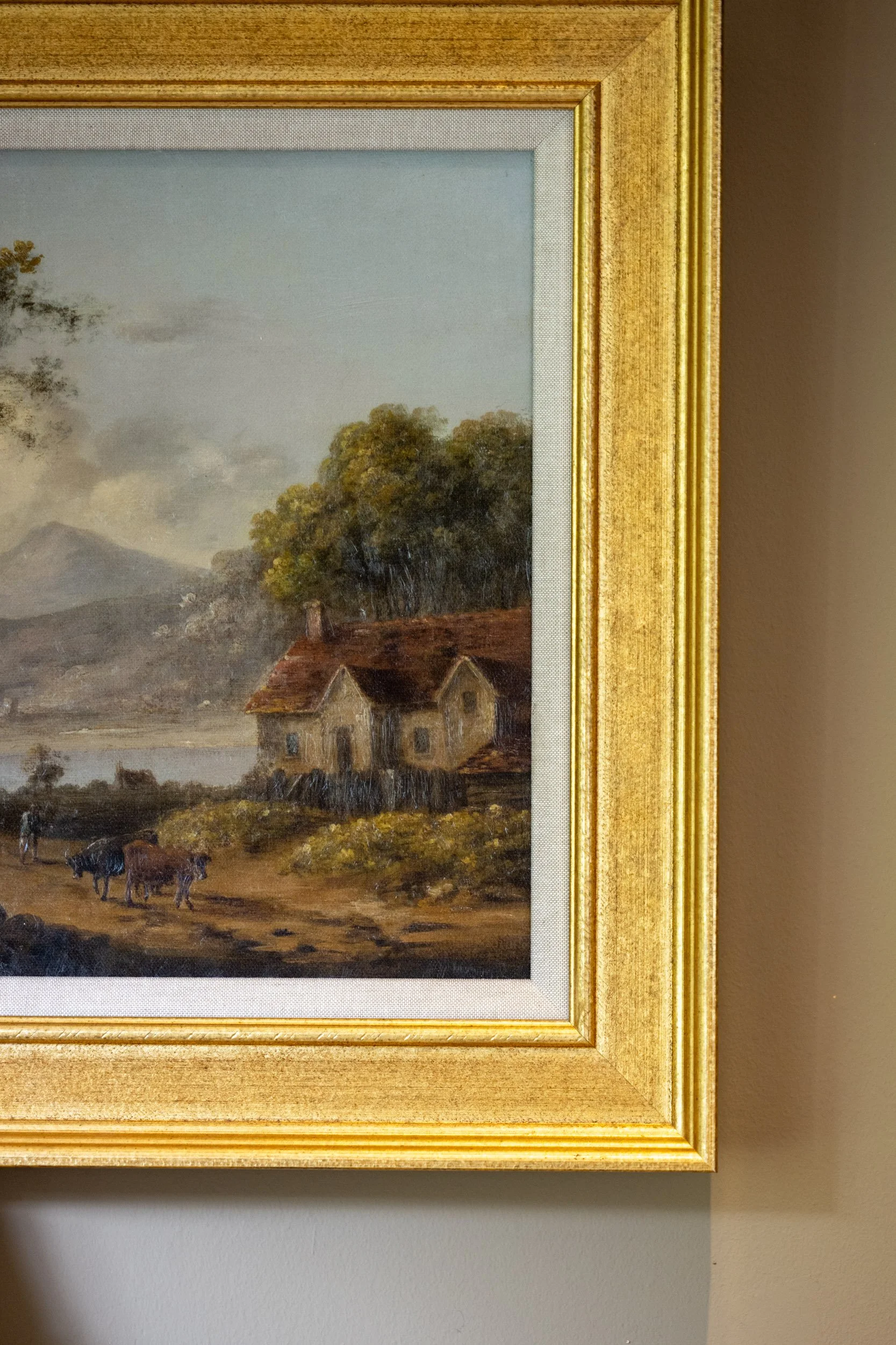 A framed landscape painting of rural scenery with trees, a house, cows, and mountains, hanging on a wall.