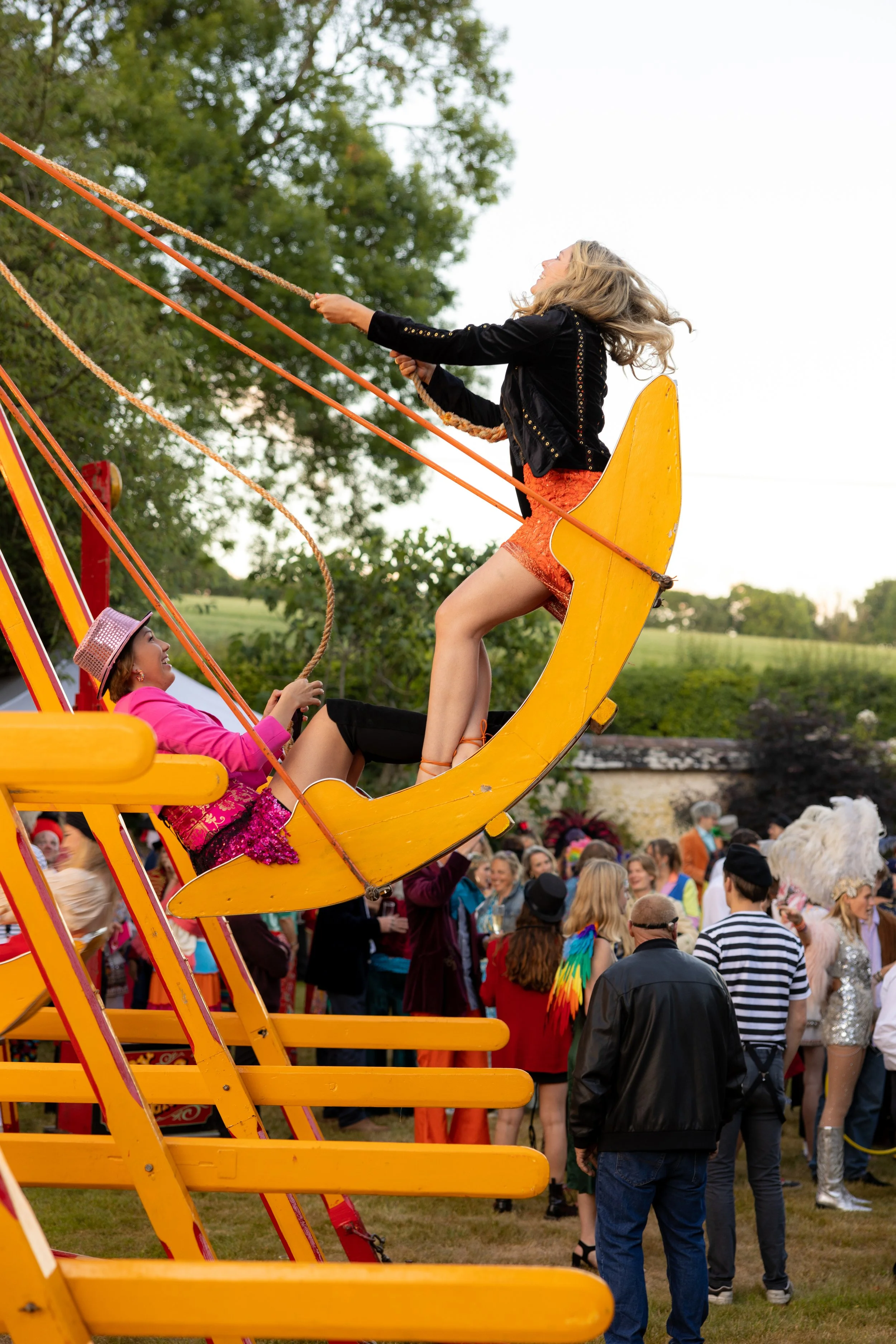 Two women on a swing ride at a fair, with one woman pushing the swing and the other sitting, surrounded by a crowd of people in colorful clothing and costumes.
