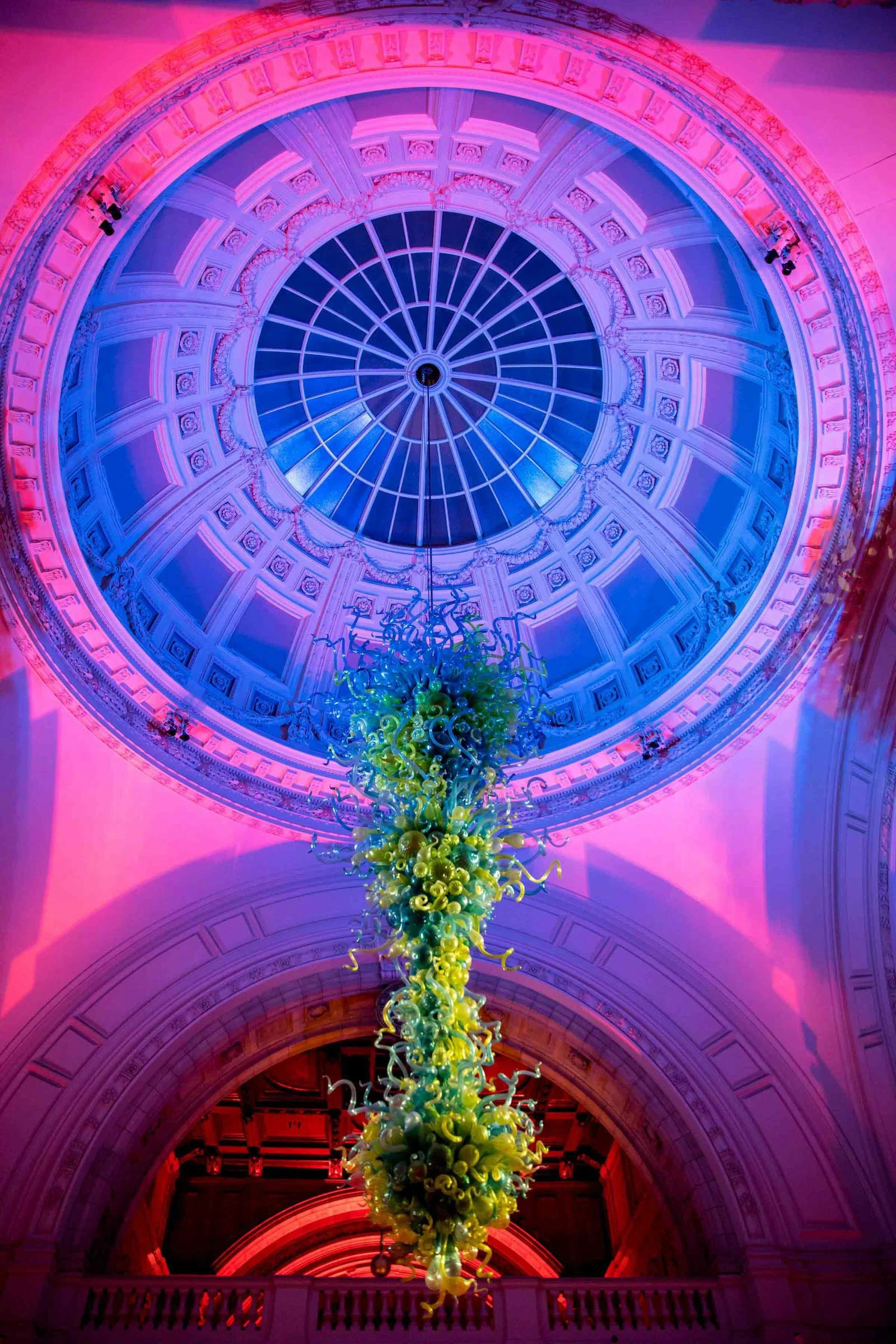 A colorful glass sculpture hangs from the ceiling of a domed architectural space with ornate detailing and a large stained glass window, illuminated with pink, blue, and purple lights.