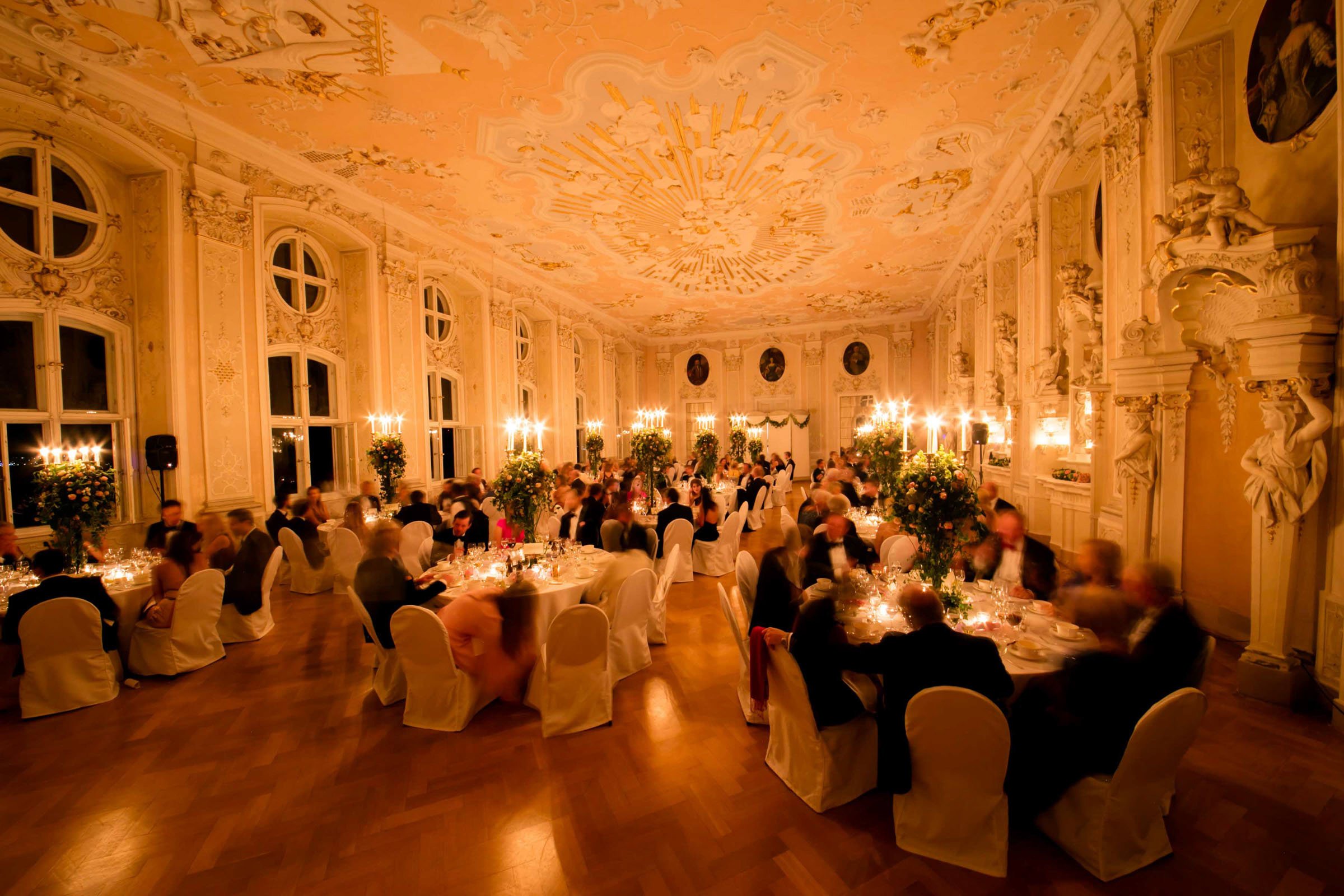 Elegant banquet hall decorated for a formal event with round tables, guests in formal attire, tall floral centerpieces, and warm lighting from chandeliers and candles.
