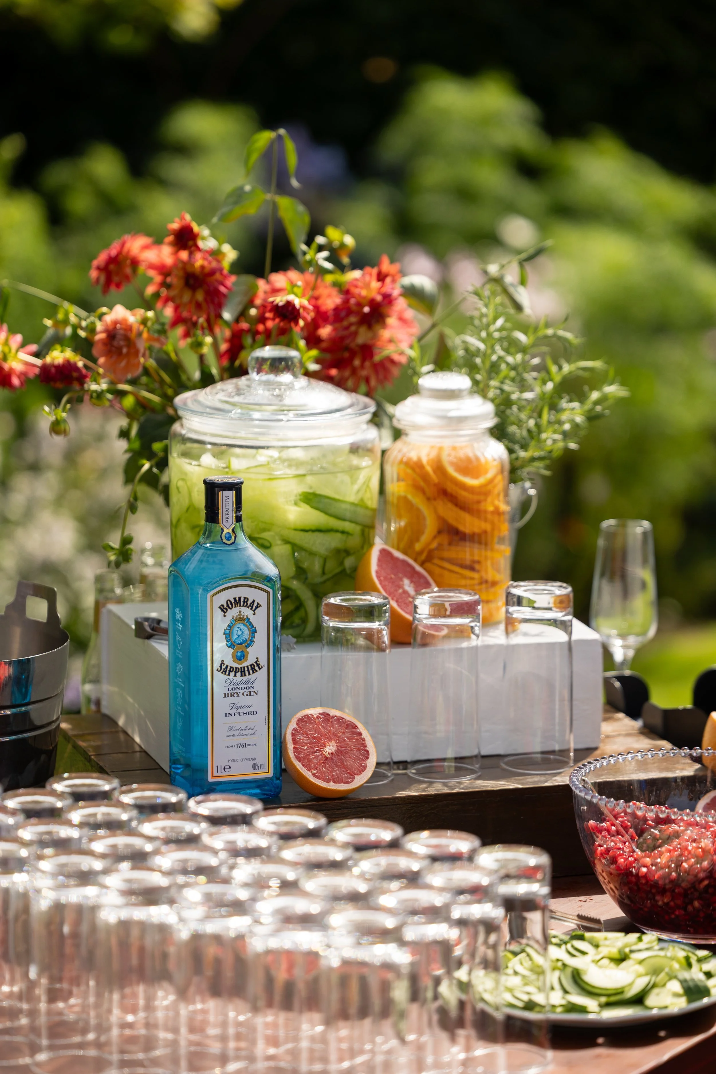 A table set outdoors with various beverages and garnishes, including a large jar of cucumber slices, a jar of orange slices, a bottle of Bombay Sapphire gin, and a halved grapefruit, surrounded by glasses and a bowl of pomegranate seeds.