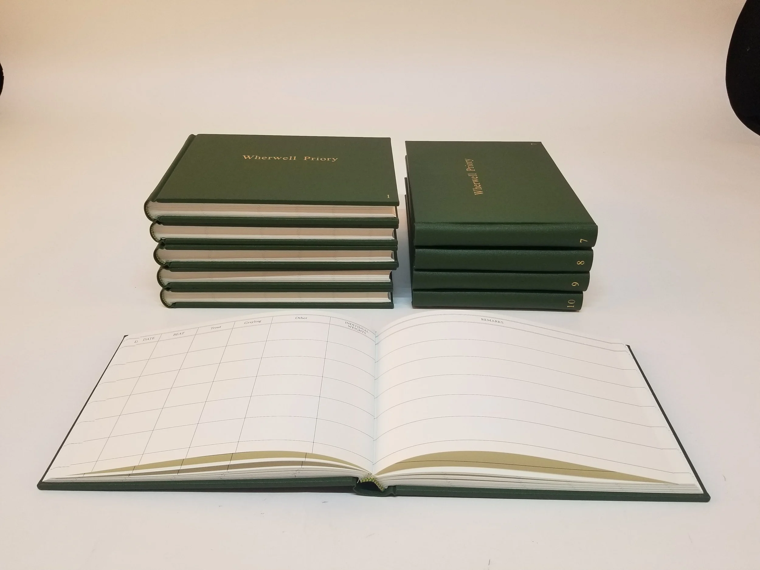 Seven green hardcover books titled "Werwell Priory" stacked on a white table, with six vertically numbered volumes and one lying flat, open planner in front.