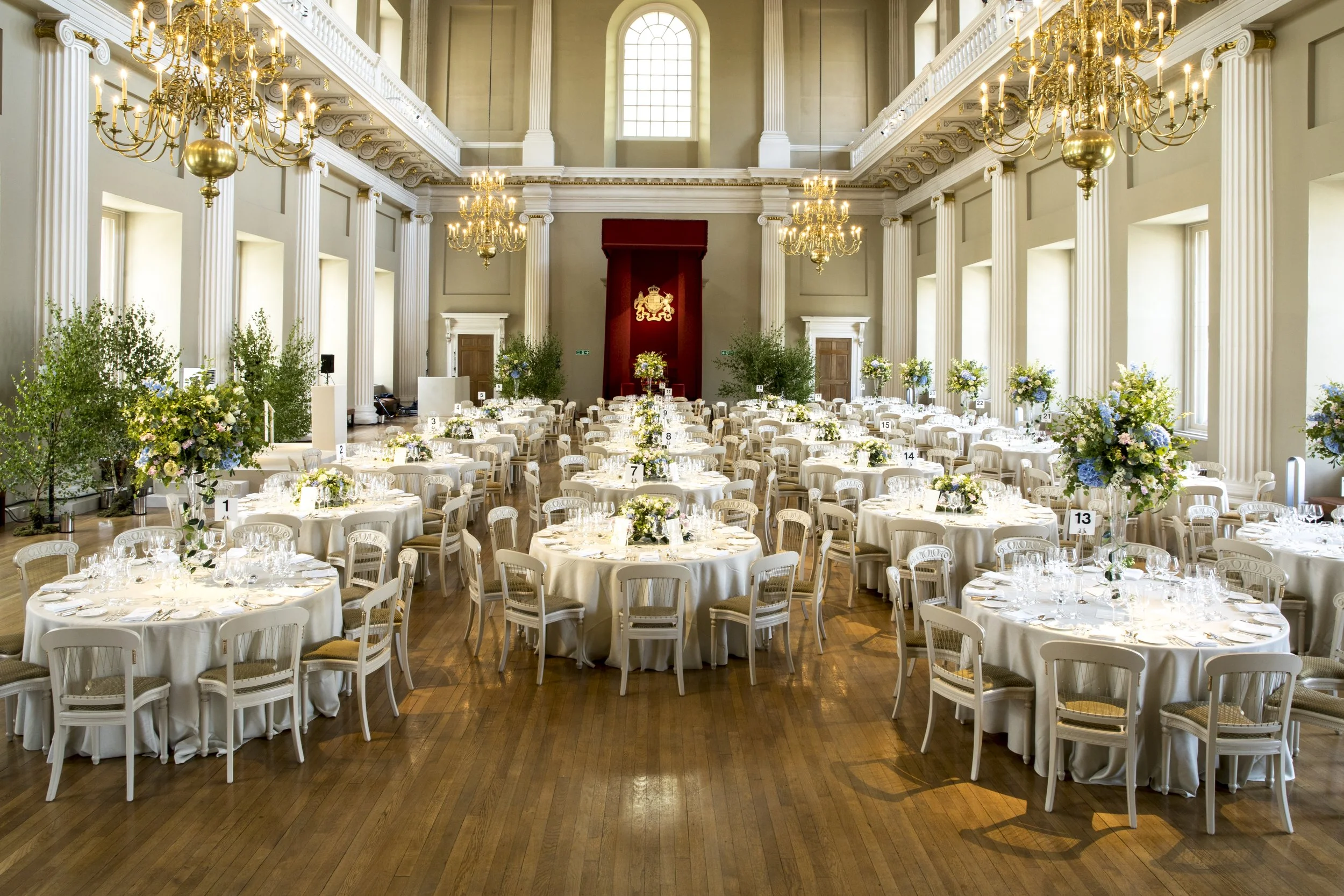 Elegant banquet hall decorated for a formal event with multiple round tables covered in white tablecloths, floral centerpieces, and surrounded by white chairs. The hall features tall windows, chandeliers, and grand columns, with a stage at the front.