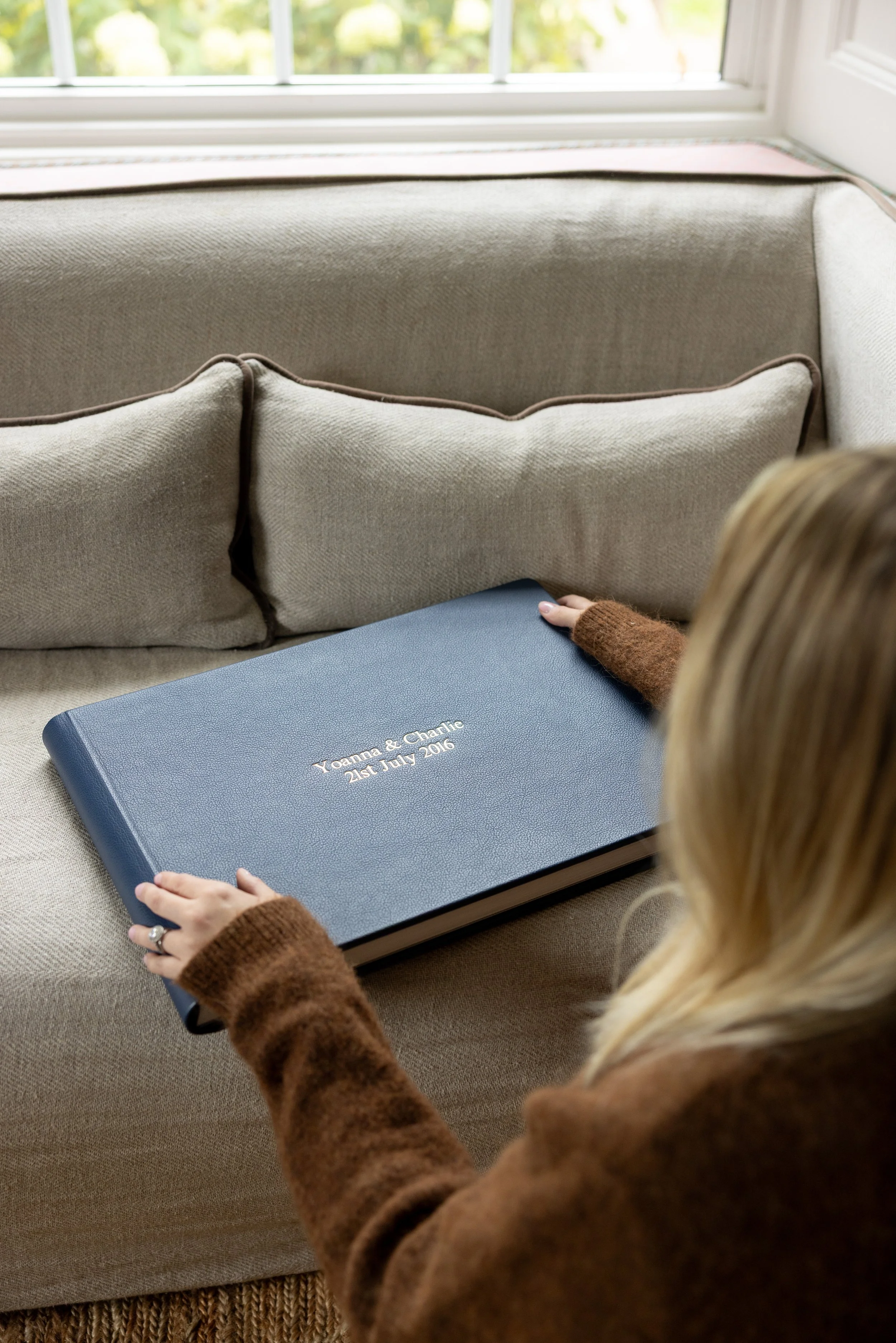 Person holding a photo album titled 'Yanna & Charlie 21st July 2016' on a beige sofa near a window.