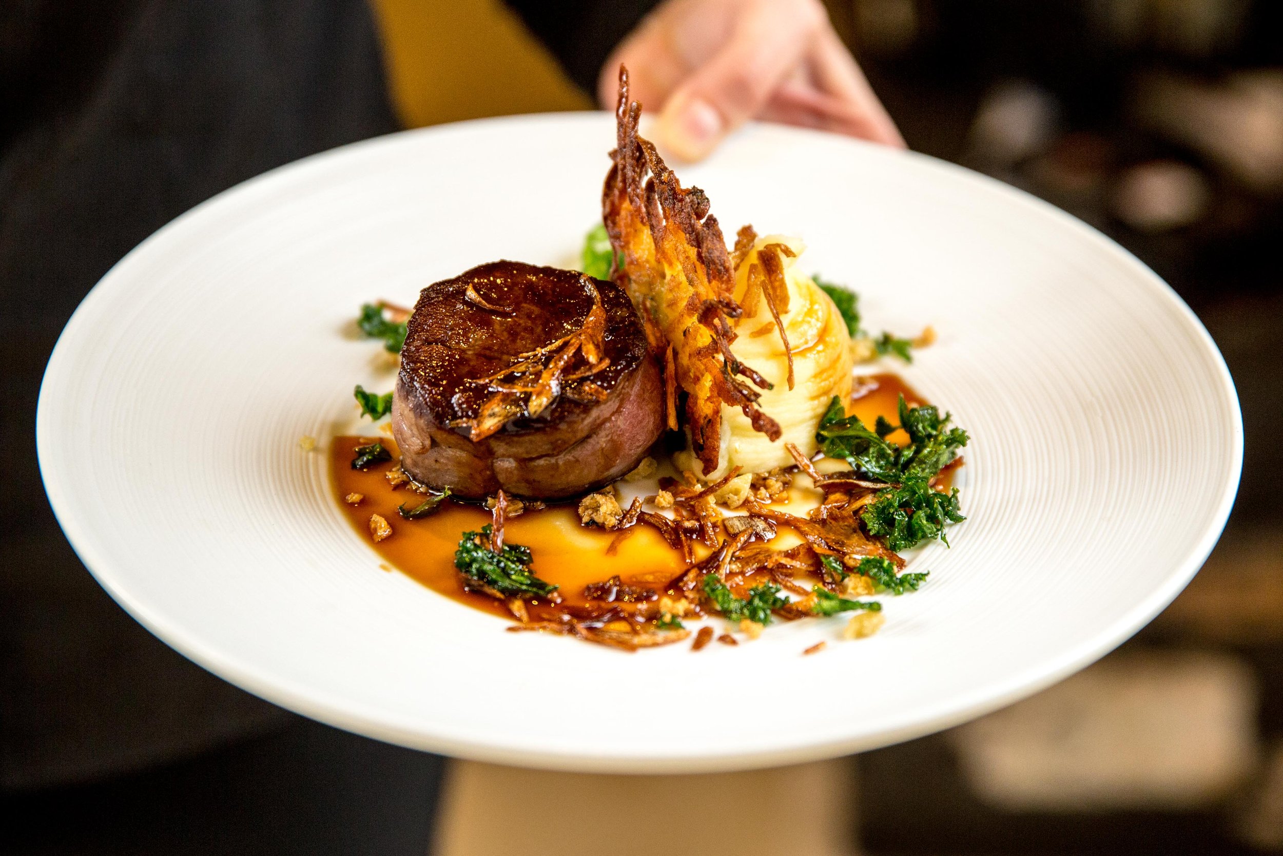 A gourmet plate featuring a seared piece of meat, mashed potatoes, crispy onion strings, and garnished greens, drizzled with gravy.