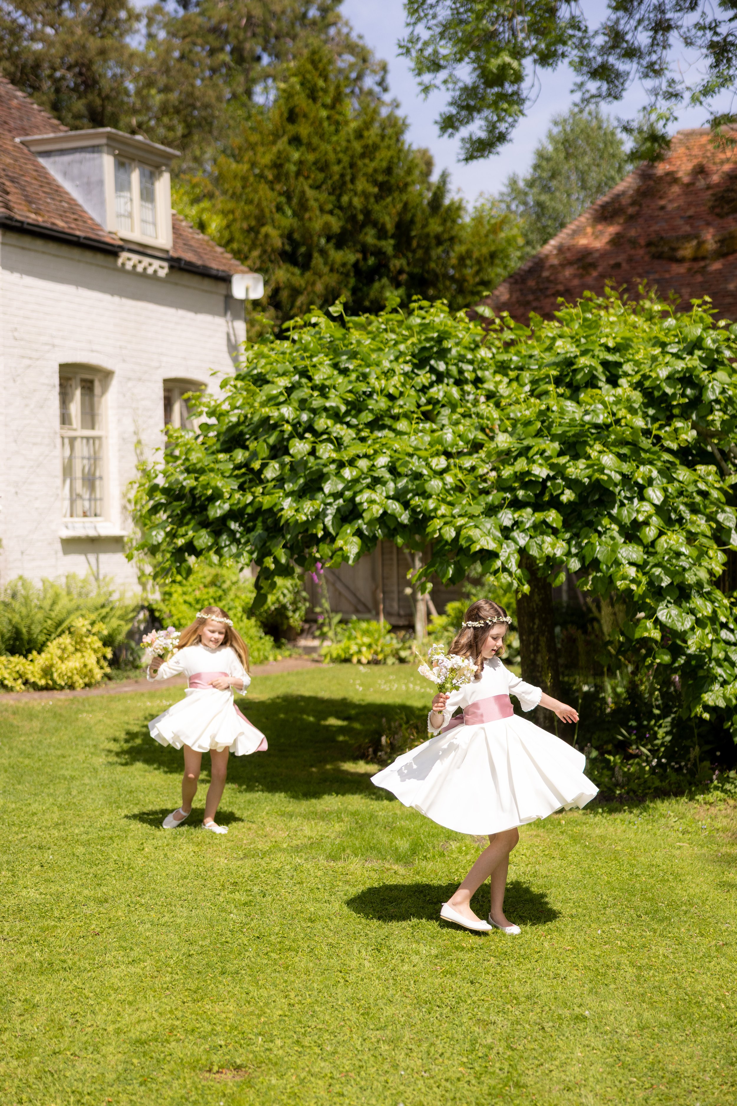 Two young girls in white dresses with pink sashes and floral headbands dancing and playing in a garden with lush green grass, bushes, and trees on a sunny day.