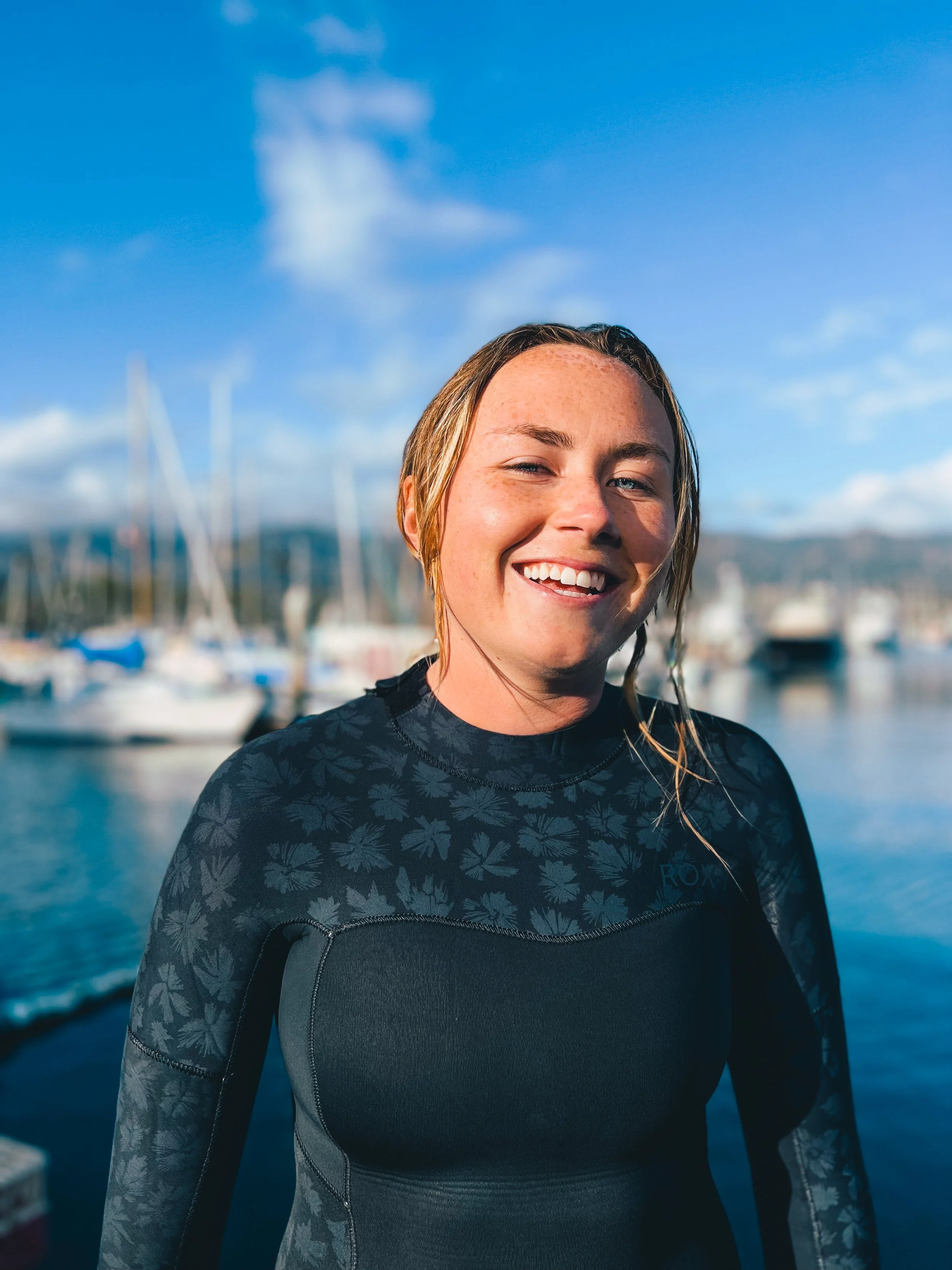 Woman in wetsuit smiling near a harbor with boats and blue sky in the background.
