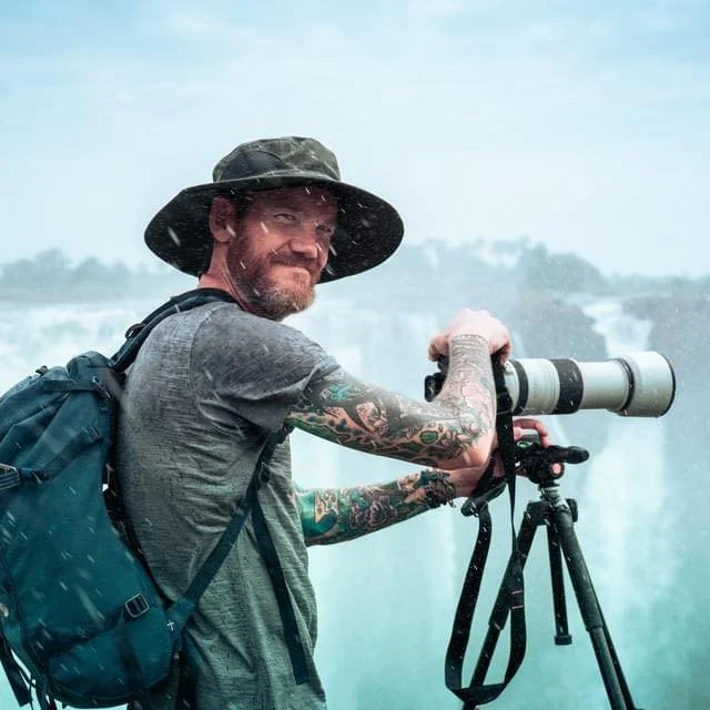 A man with tattoos and a hat using a camera with a large telephoto lens on a tripod, at a scenic outdoor location with waterfalls and mist.
