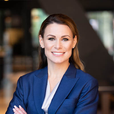 A professional woman with long brown hair, wearing a blue blazer and white blouse, smiling confidently in an indoor setting.