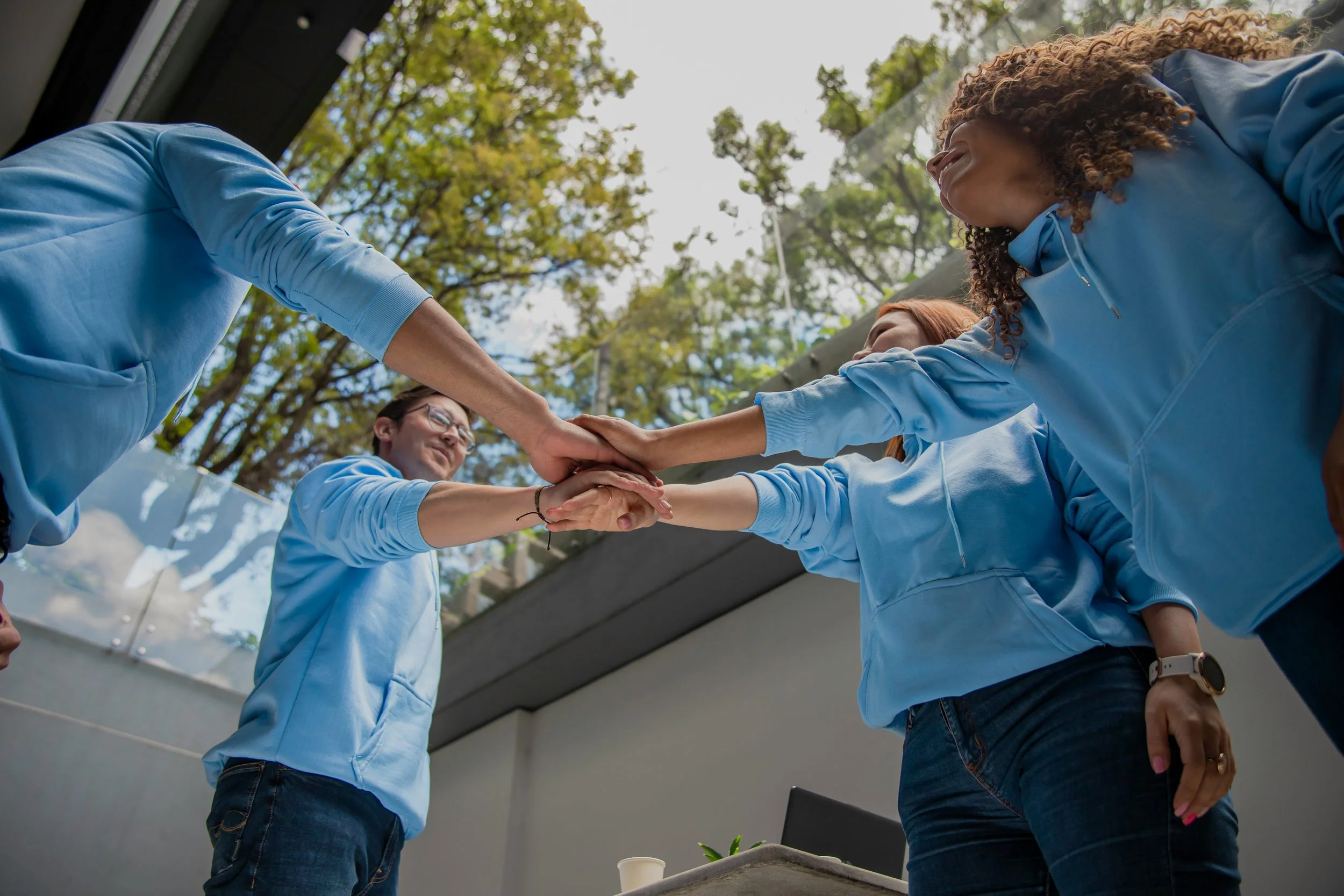 Group of diverse young people wearing blue hoodies, standing in a circle outdoors, putting their hands together in the center for a team hug or cheer, with trees and blue sky in the background.