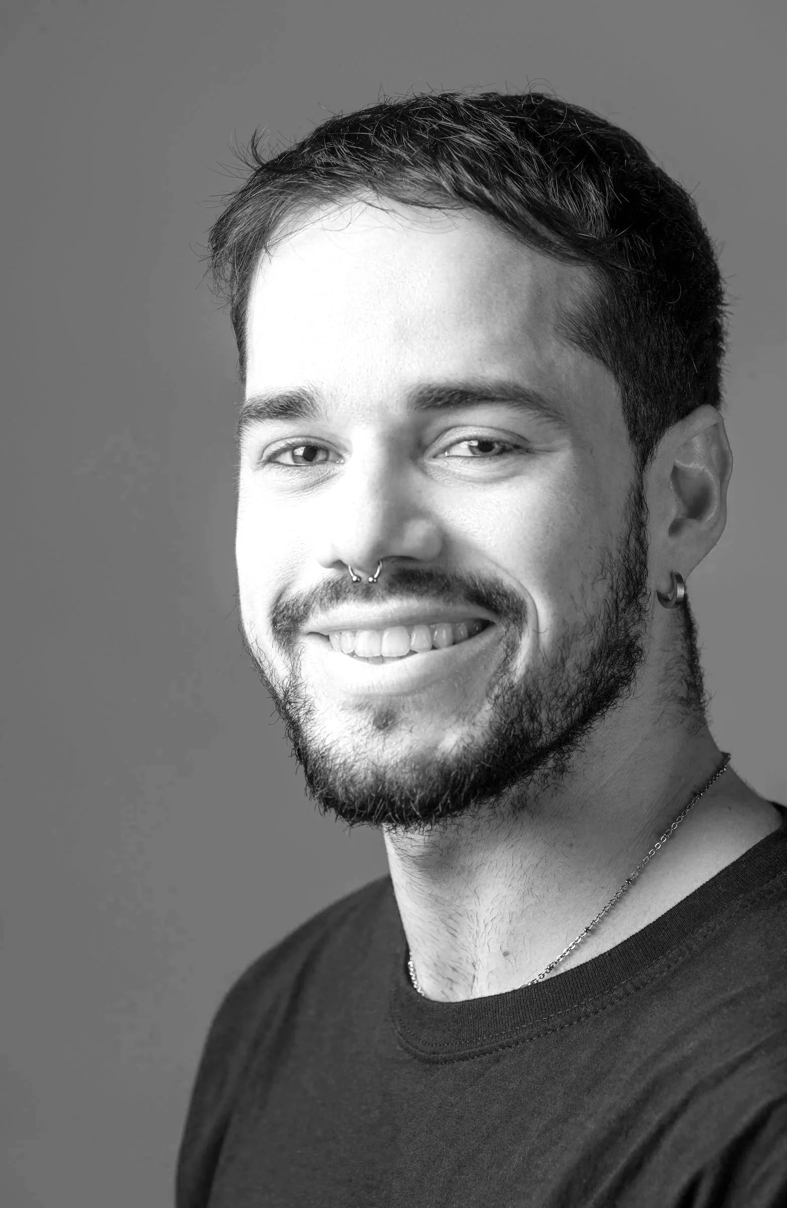 Close-up portrait of a smiling man with short dark hair, blue eyes, a septum piercing, a beard, and wearing earrings, a gold chain, and a black shirt against a plain brown background.