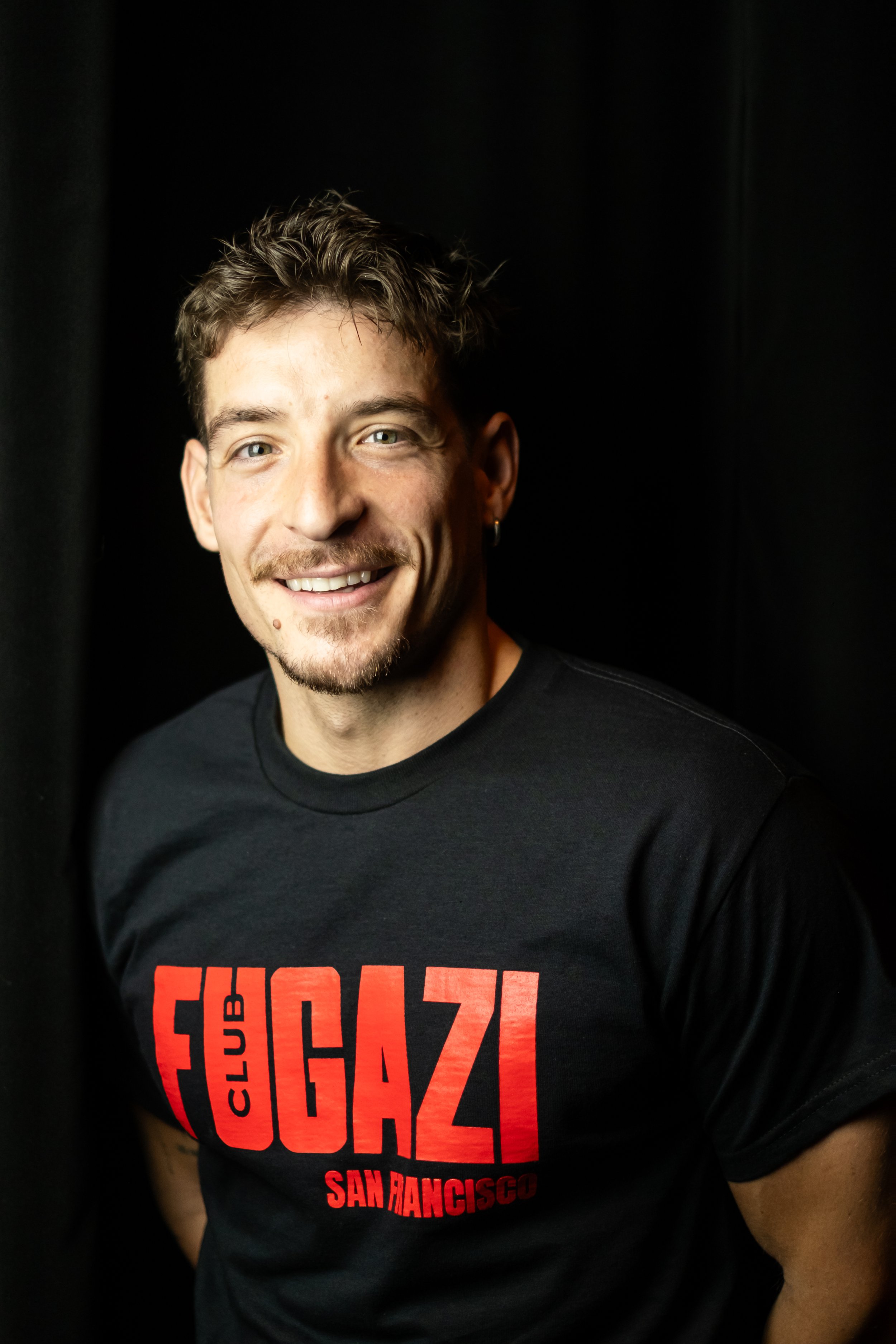 A young man with short dark hair smiling, wearing a black and white polka dot shirt over a white t-shirt, standing in front of dark curtains with a pink light highlighting his face.