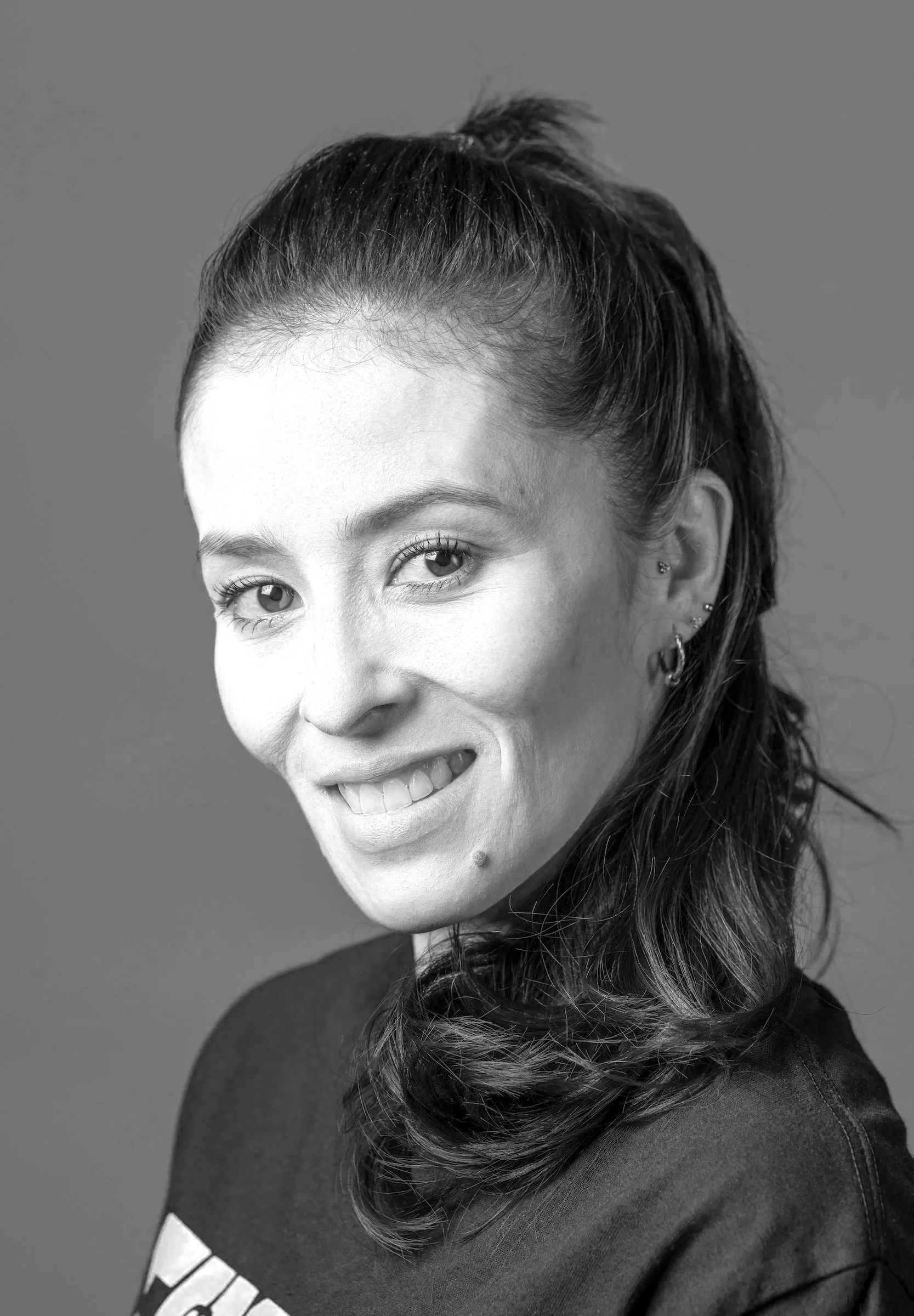 A smiling woman with shoulder-length dark brown hair, blue eyes, wearing earrings, and a dark t-shirt, posing against a plain brown background.