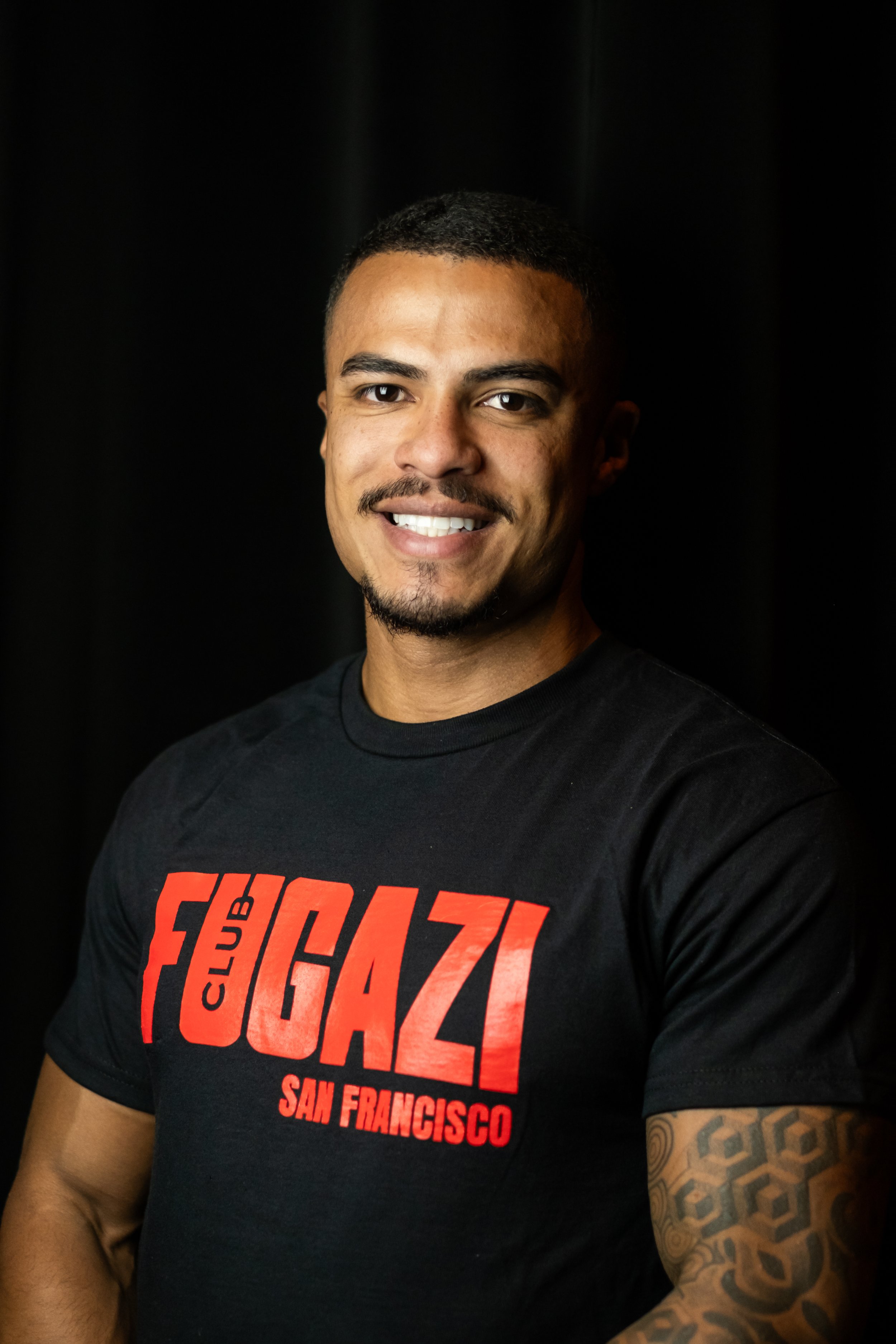 A smiling man with a beard and mustache wearing a black t-shirt with red and white text that reads "Club GAZ San Francisco" against a black background.