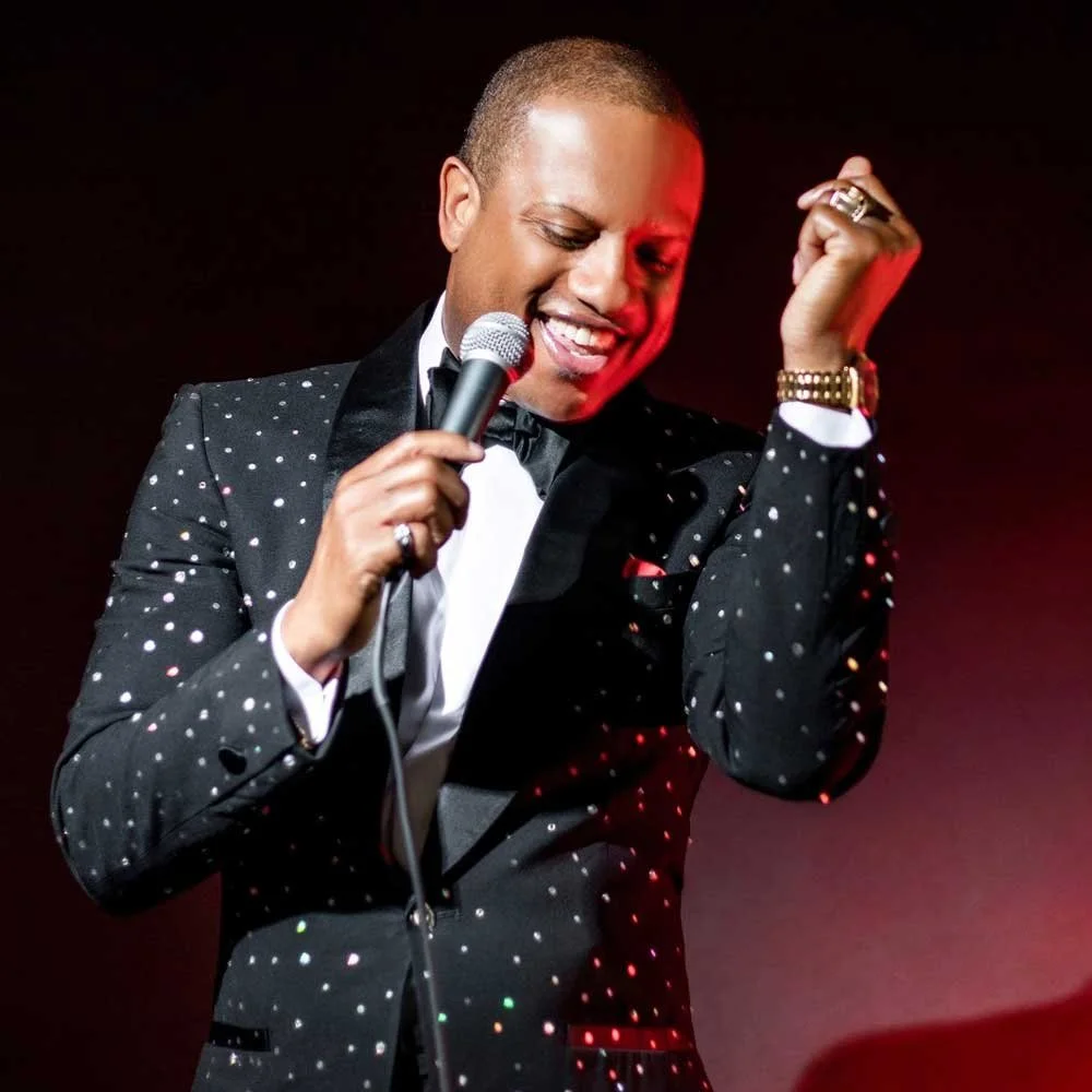 A man in a black tuxedo with white dots, holding a microphone, smiling, with a dark background.