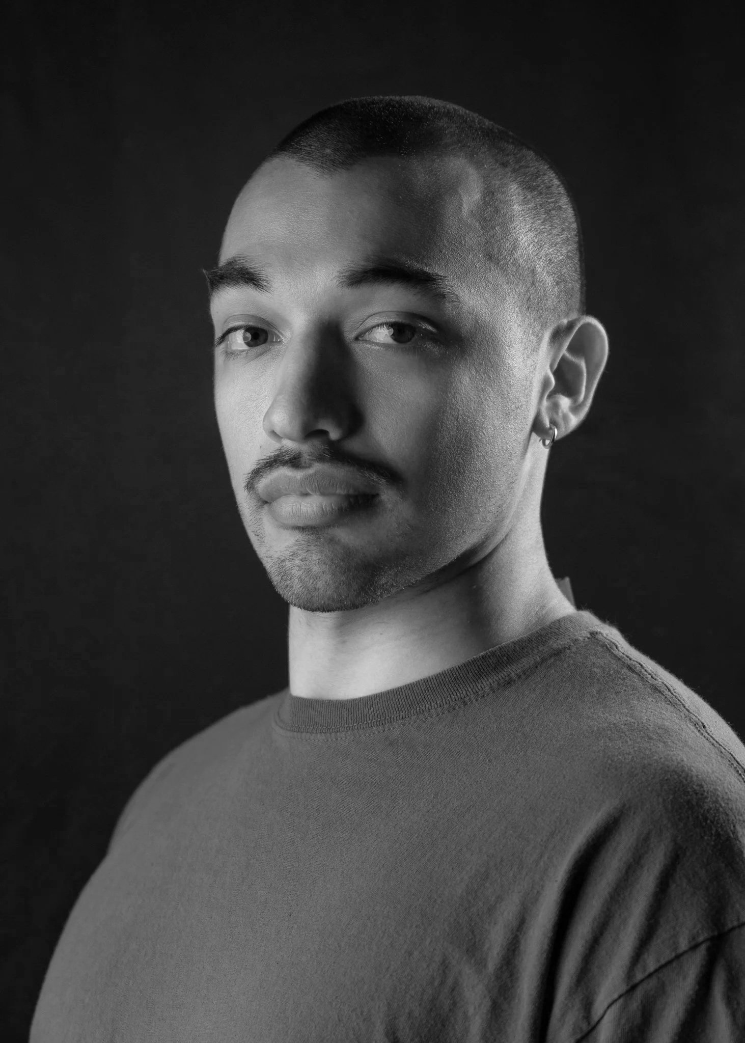 Portrait of a young man with a short haircut, light facial hair, and small hoop earring, wearing a gray T-shirt against a black background.