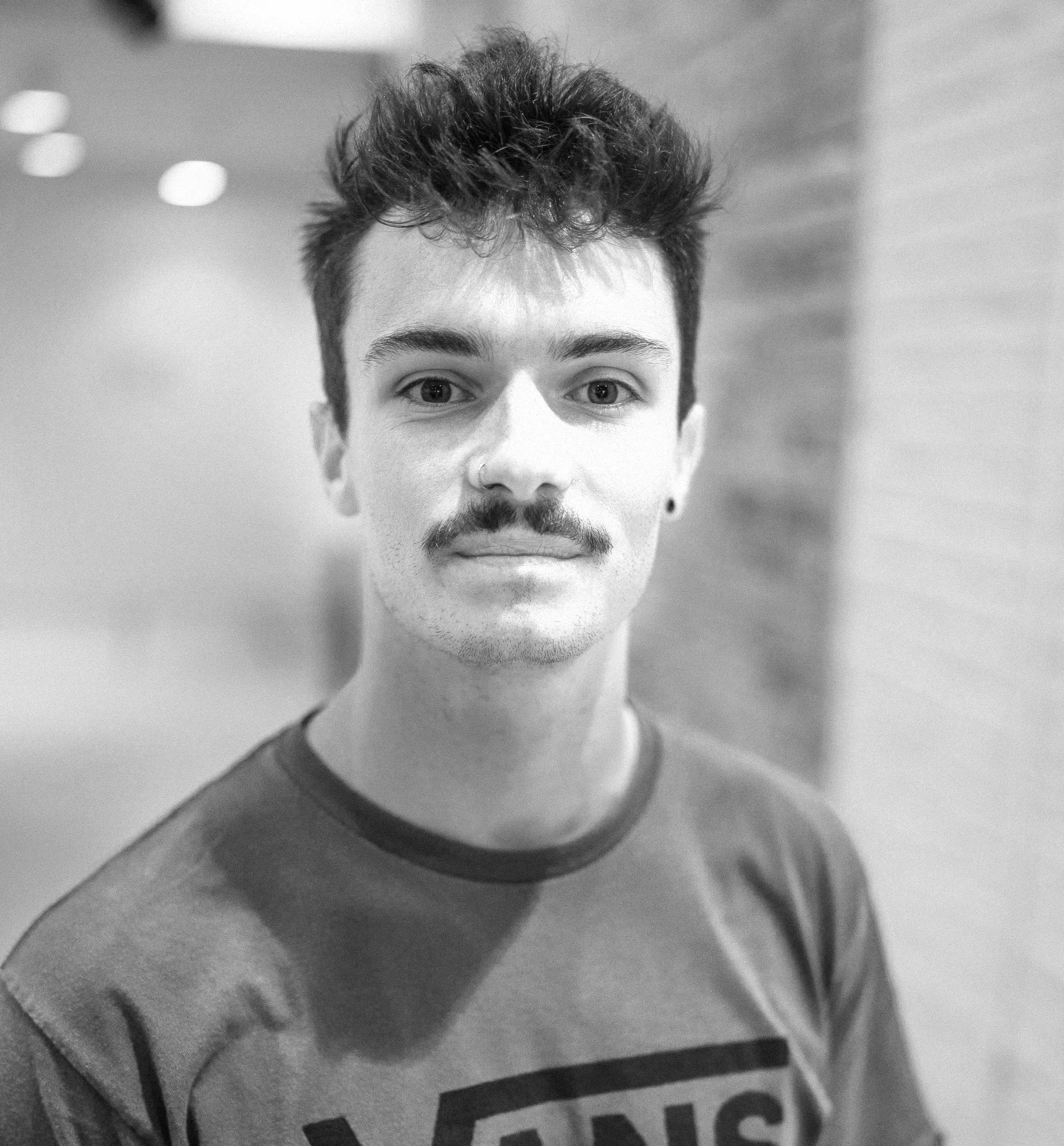 A young man with dark curly hair, a mustache, goatee, and nose piercing, wearing a red T-shirt, standing indoors in front of a blurred background with a brick wall and white surface.