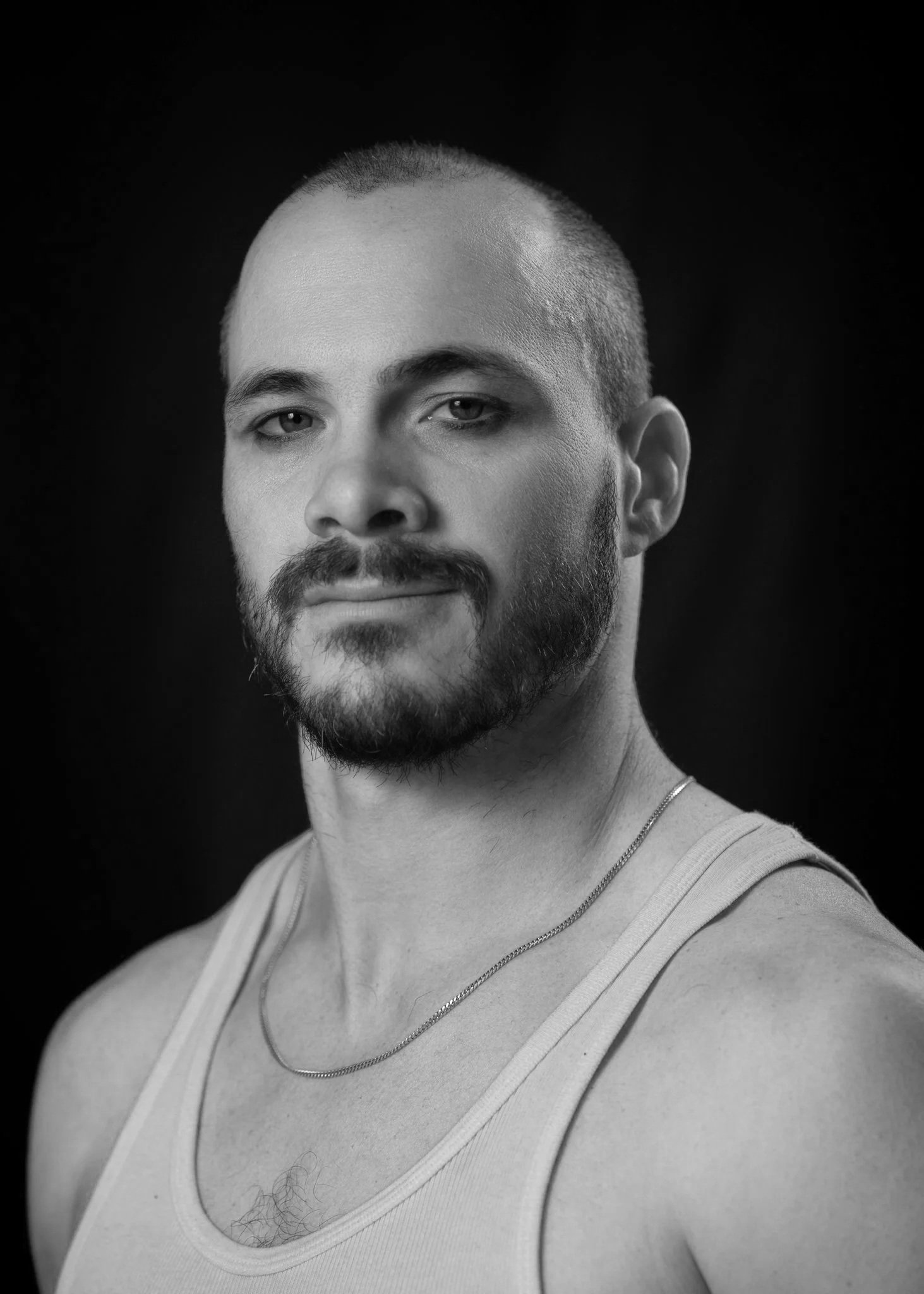 Close-up portrait of a man with a beard, mustache, and short hair, wearing a white tank top and a gold chain necklace against a black background.