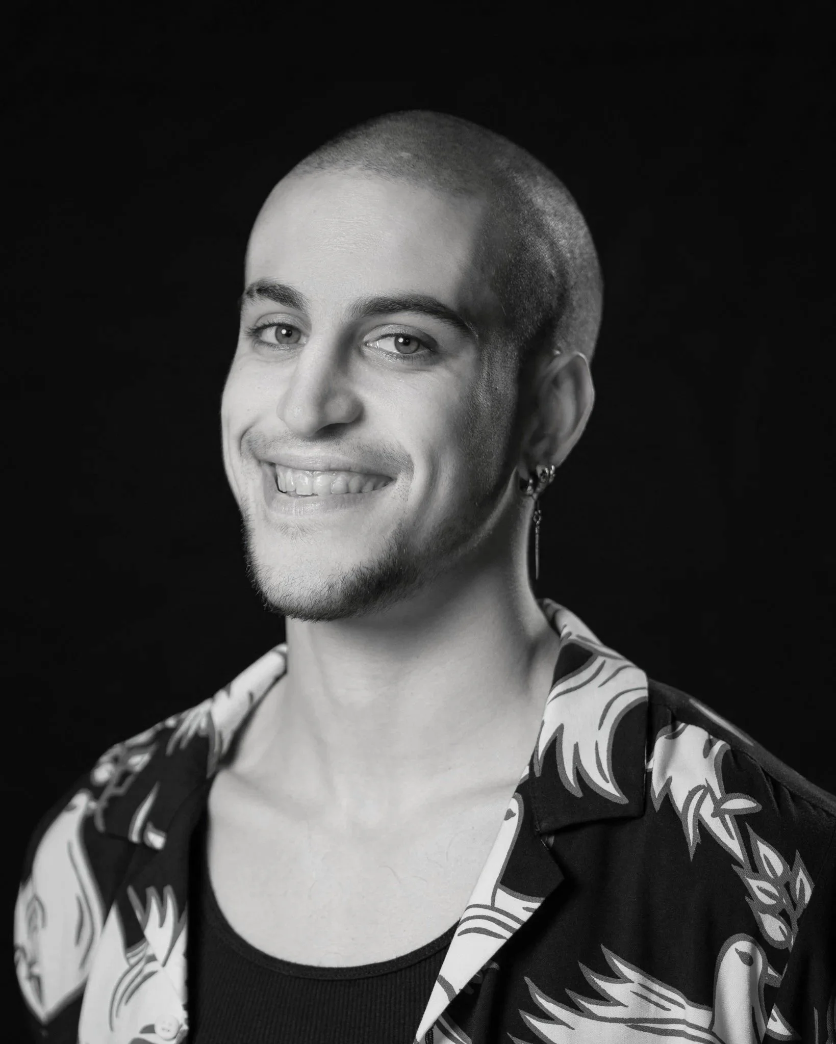 Portrait of a smiling young man with a shaved head, earrings, and a tattoo visible on his neck, wearing a patterned shirt against a black background.