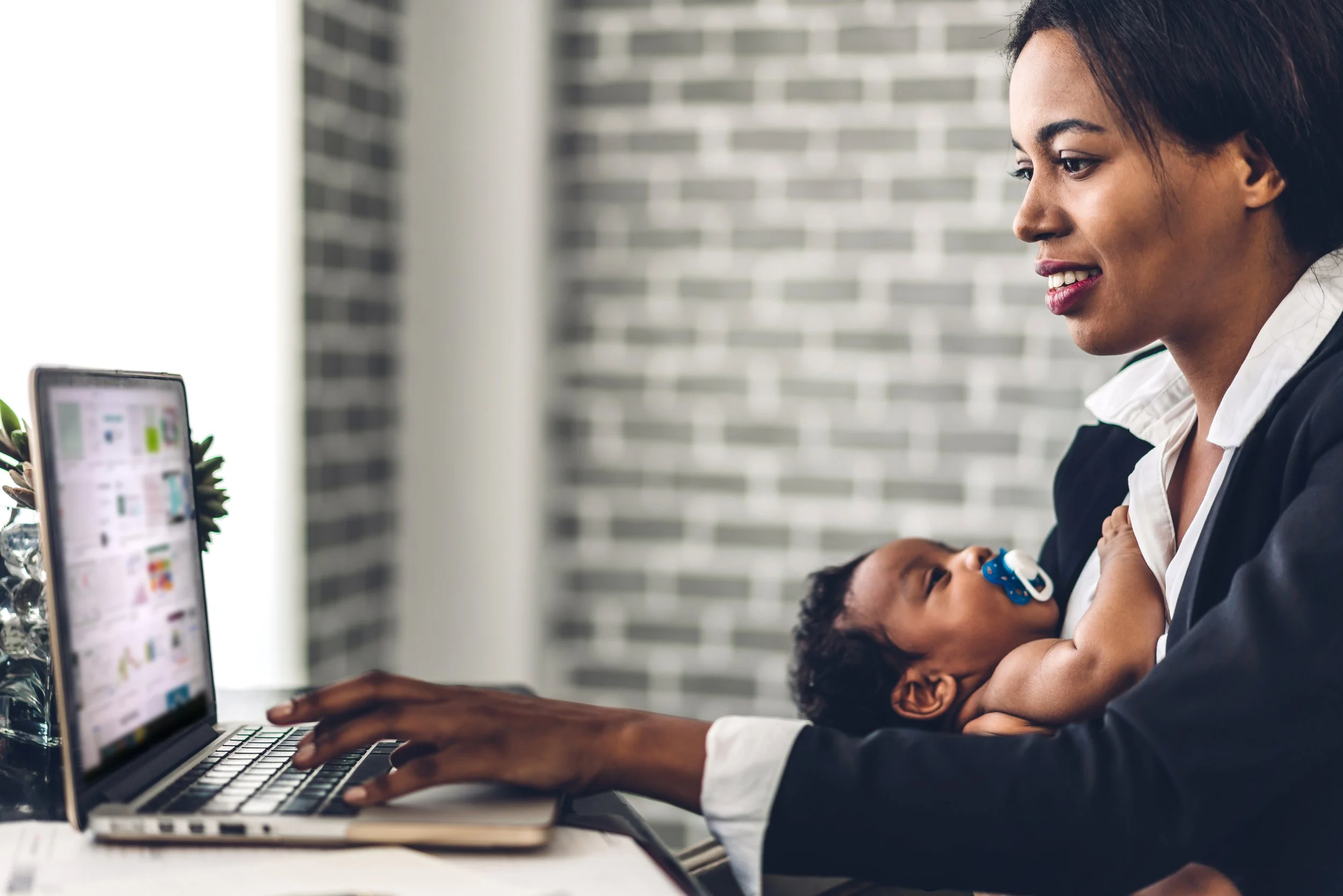 A woman sits at a desk with a laptop, breastfeeding a baby who has a pacifier, in a room with a brick wall background.
